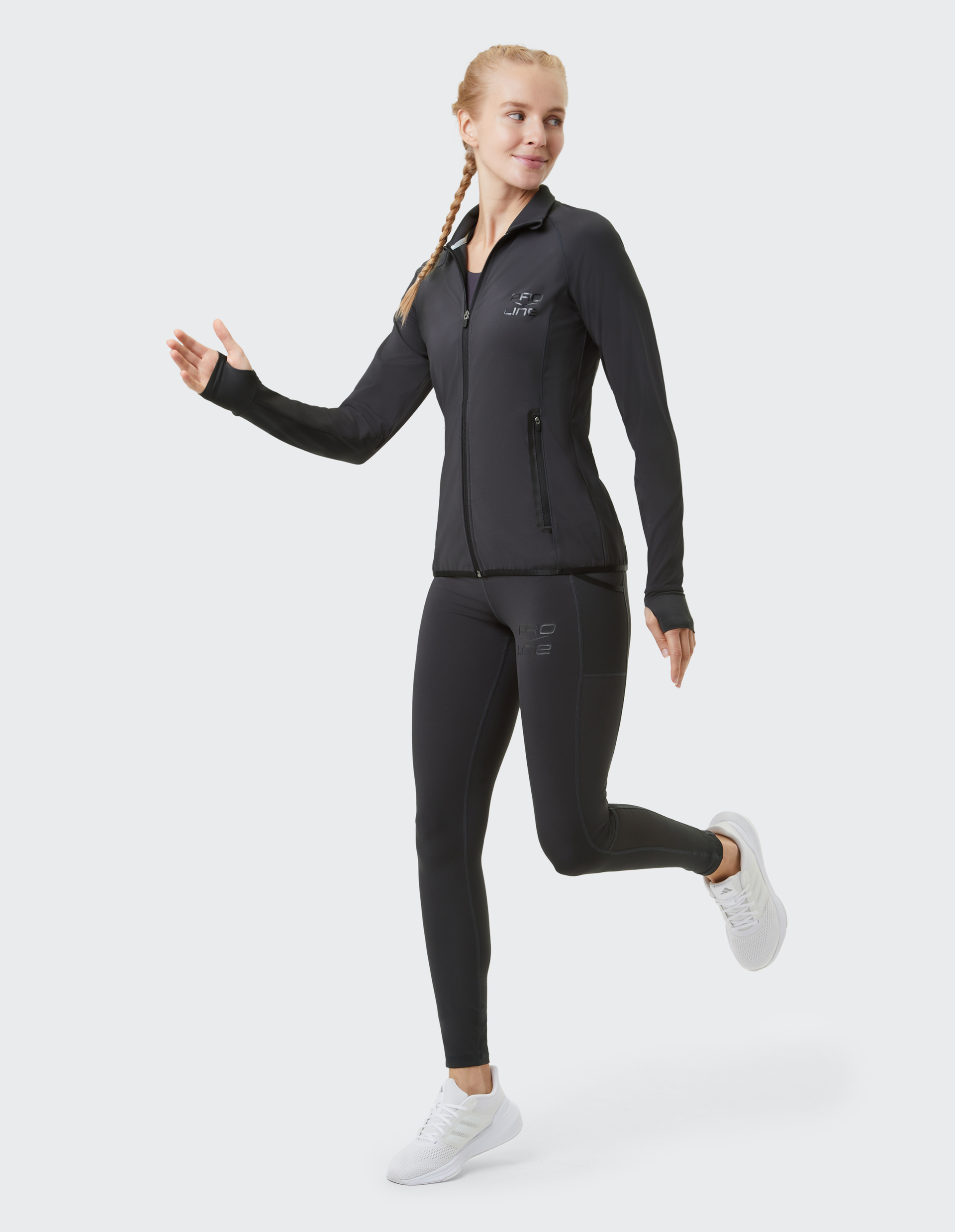 Frau in schwarzer Sportkleidung mit Zopf joggt lächelnd, trägt weiße Turnschuhe und hebt das linke Bein vor neutralem Hintergrund.