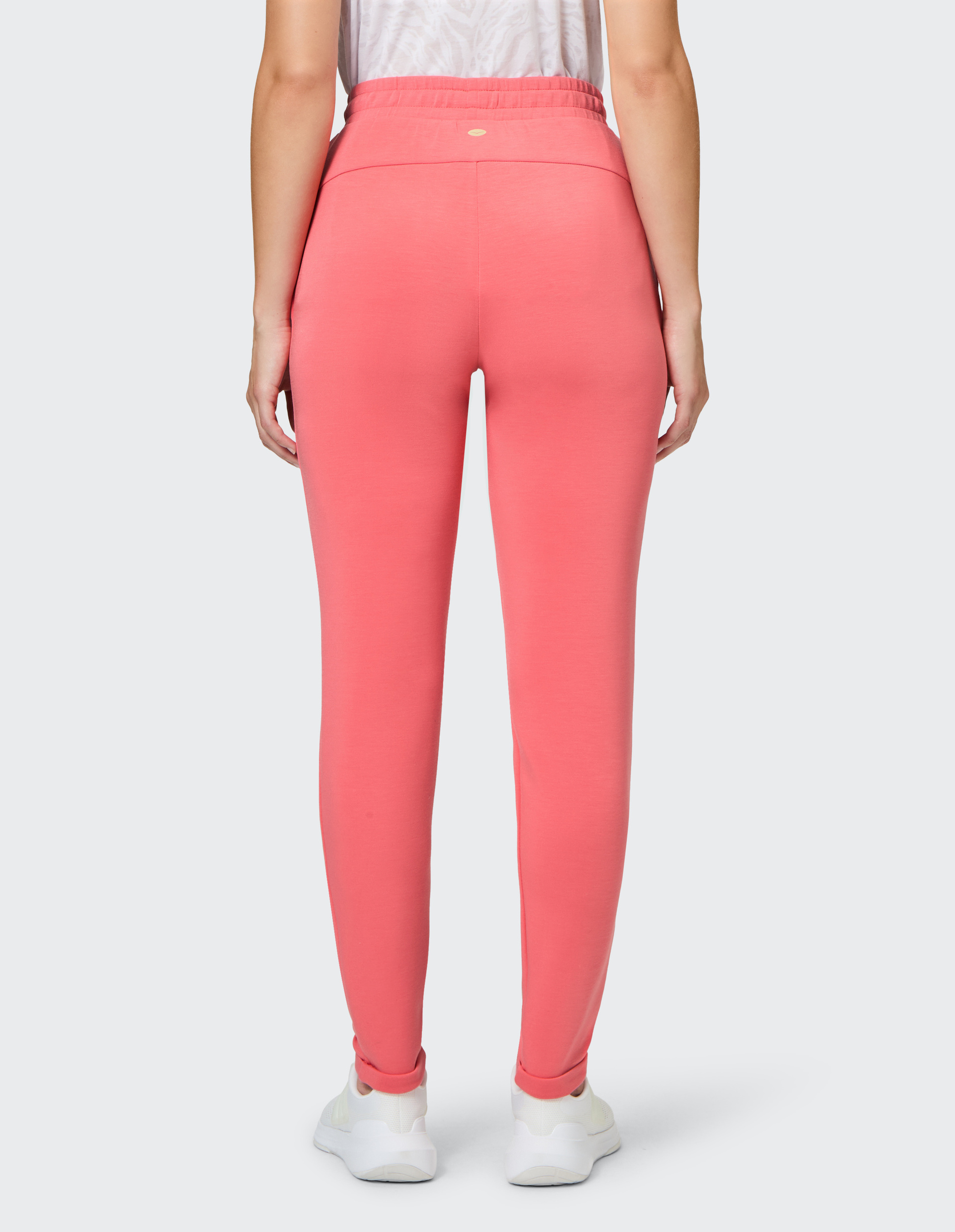 Person von hinten in korallfarbener Leggings und weißem T-Shirt, trägt weiße Sneaker, fotografiert gegen hellen Hintergrund.