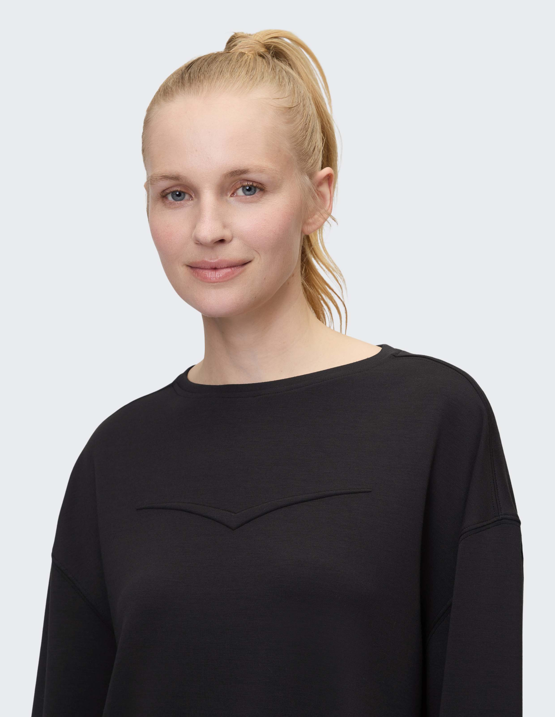 Eine lächelnde Person mit blondem Pferdeschwanz trägt ein schwarzes Langarmshirt und blickt direkt in die Kamera vor grauem Hintergrund.