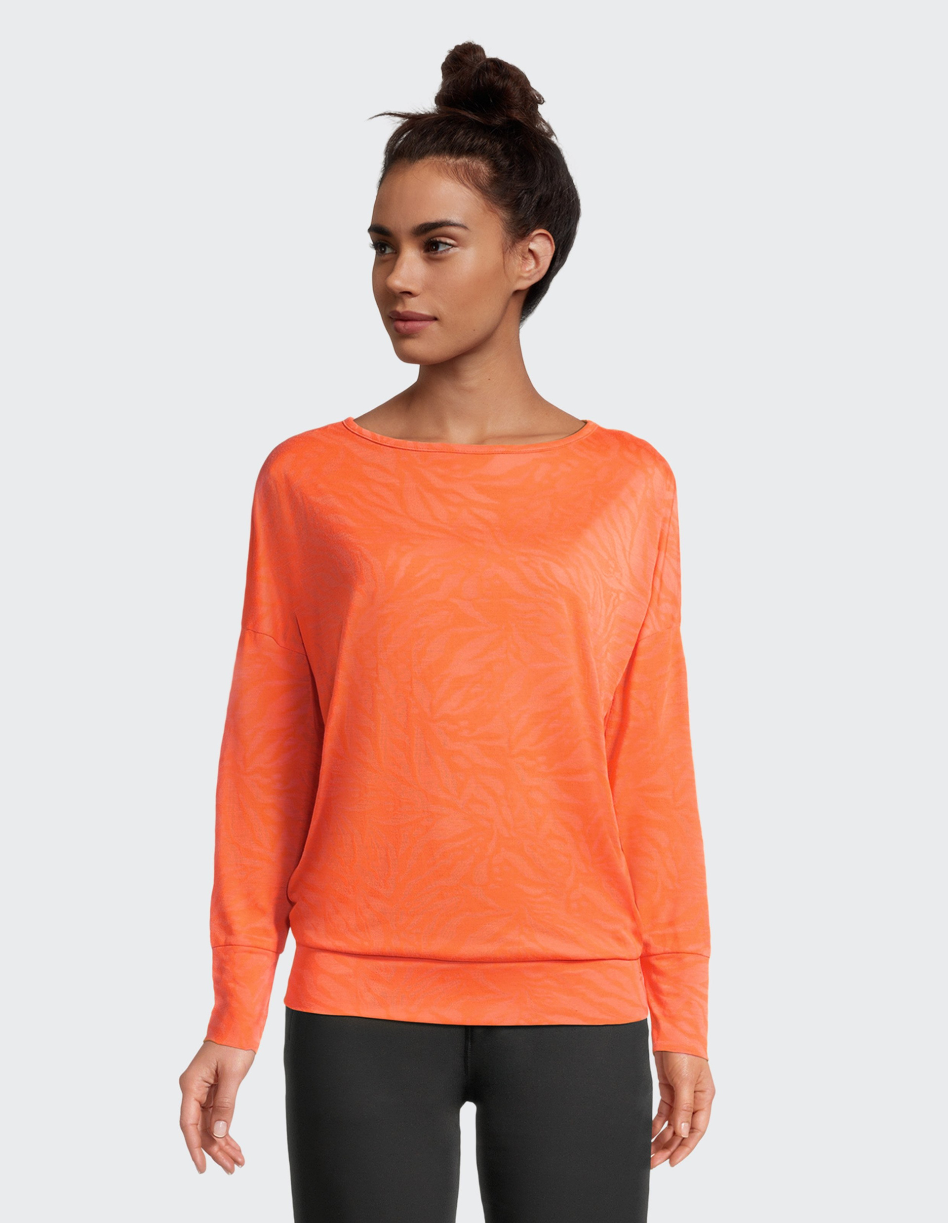 Frau trägt leuchtend orangefarbenen Pullover mit dezentem Muster und schwarzen Hosen, Haare in lockerem Dutt, vor grauem Hintergrund.