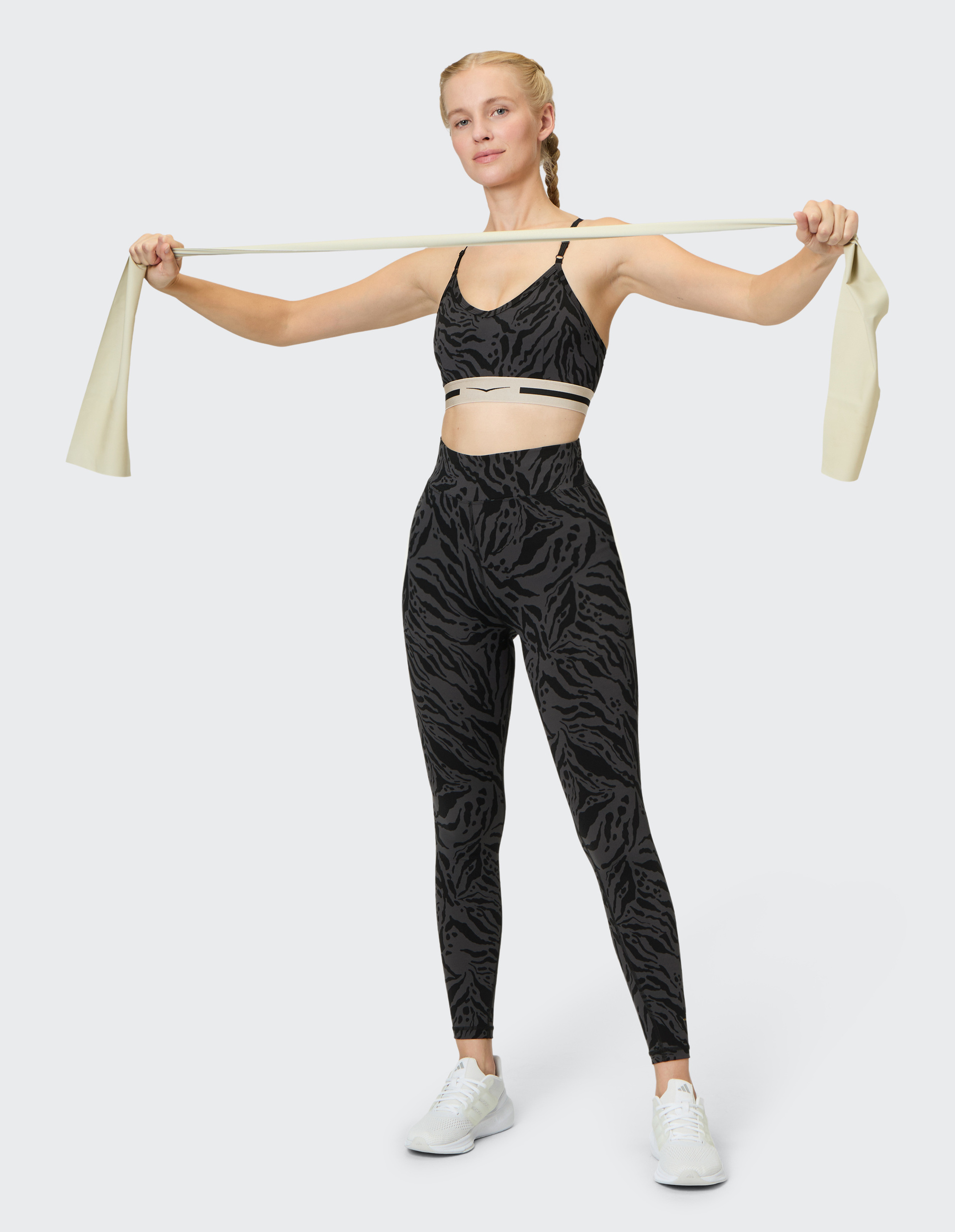 Frau in gemustertem Sportoutfit dehnt mit hellen Widerstandsband, Gesicht selbstbewusst, langer Zopf, trägt weiße Sportschuhe.