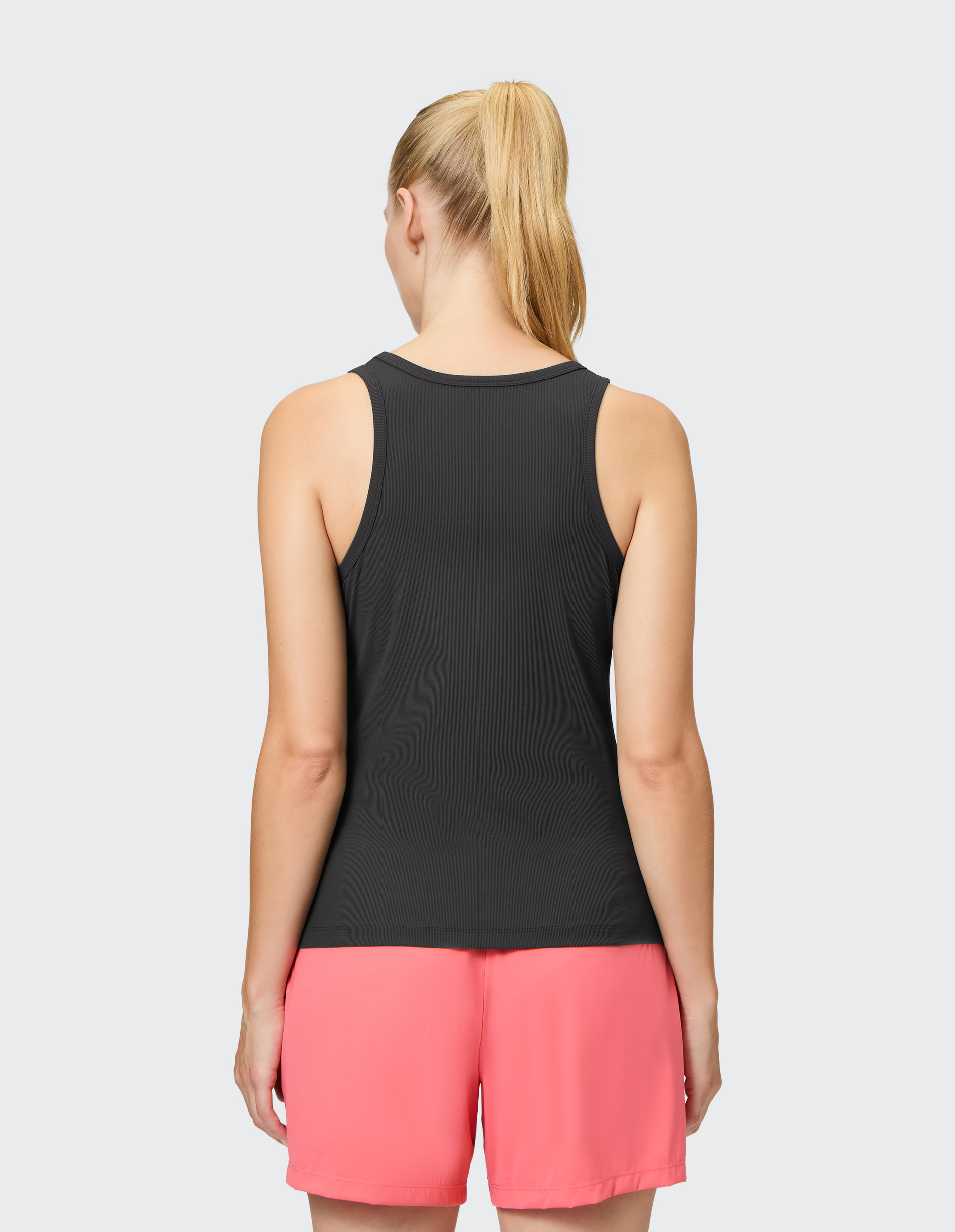 Frau mit blondem Pferdeschwanz von hinten, trägt schwarzes Tanktop und pinke Shorts, steht vor hellgrauem Hintergrund.