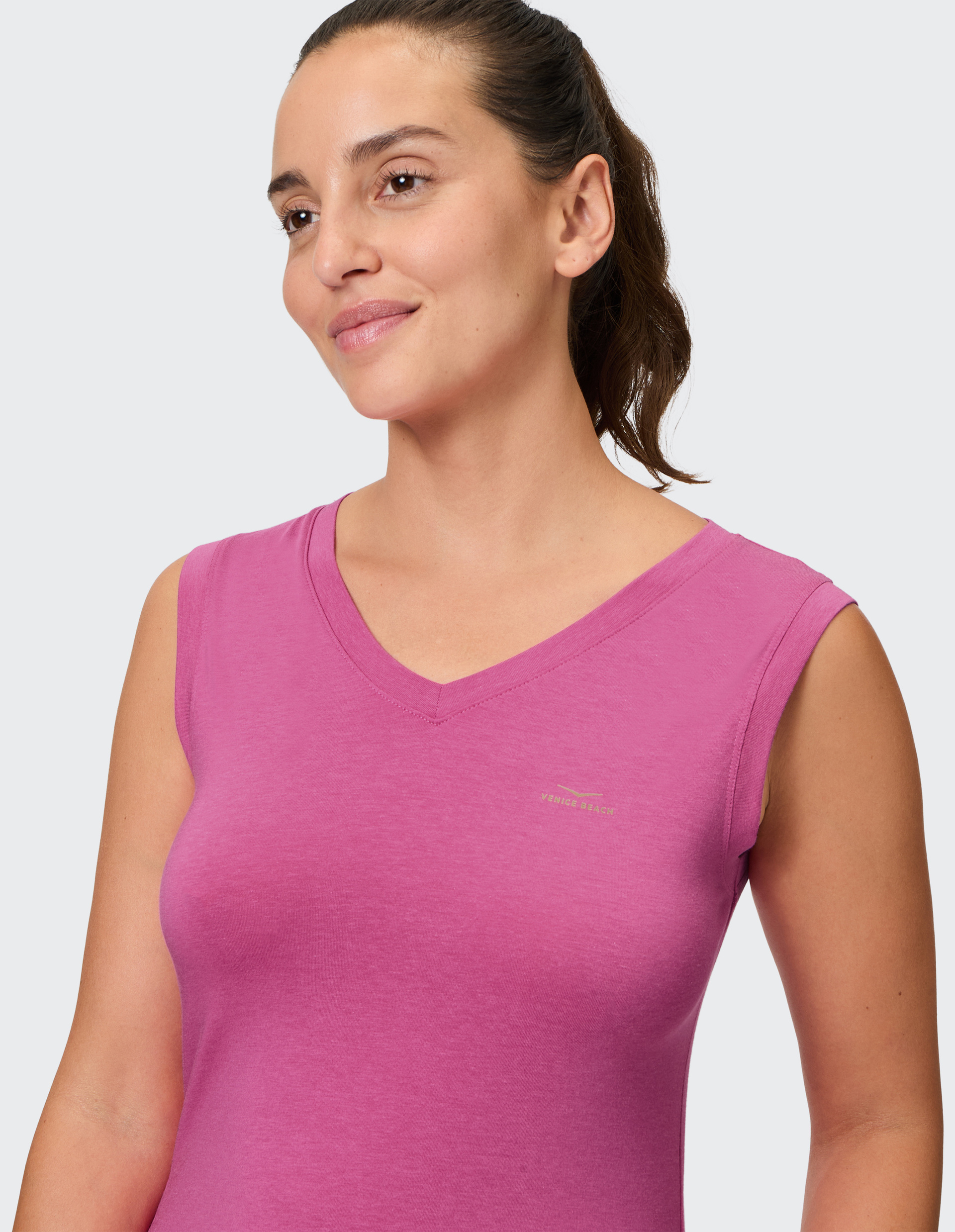 Frau mit seitlich gebundenem Haar trägt ein pinkes ärmelloses Oberteil und lächelt leicht vor hellem Hintergrund.