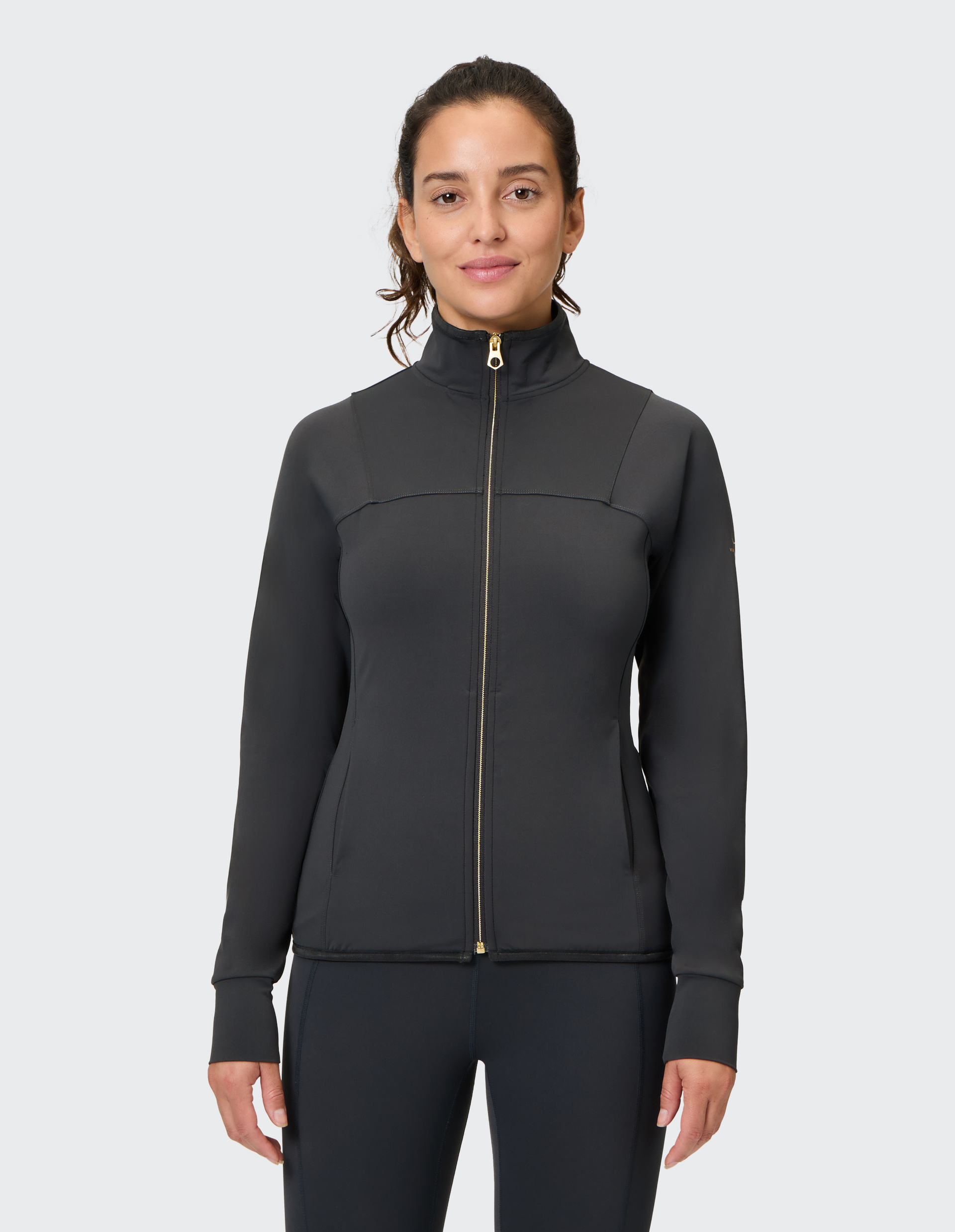 Frau in schwarzer Sportjacke mit Reißverschluss und hohem Kragen, lächelnd, vor einem hellgrauen Hintergrund.