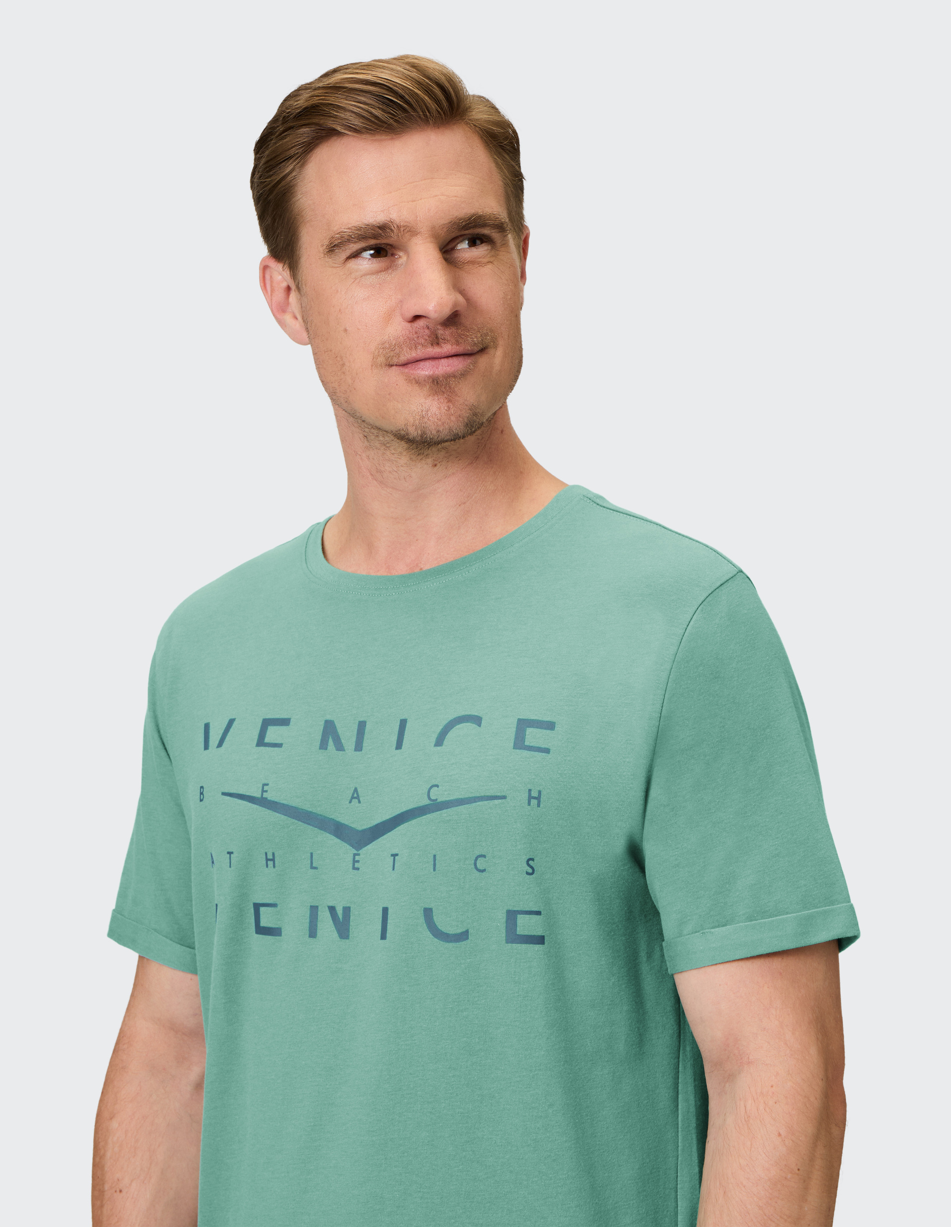 Junger Mann mit kurzem Haar trägt ein grünes T-Shirt mit blauem "Venice Beach Athletics"-Aufdruck und schaut zur Seite.