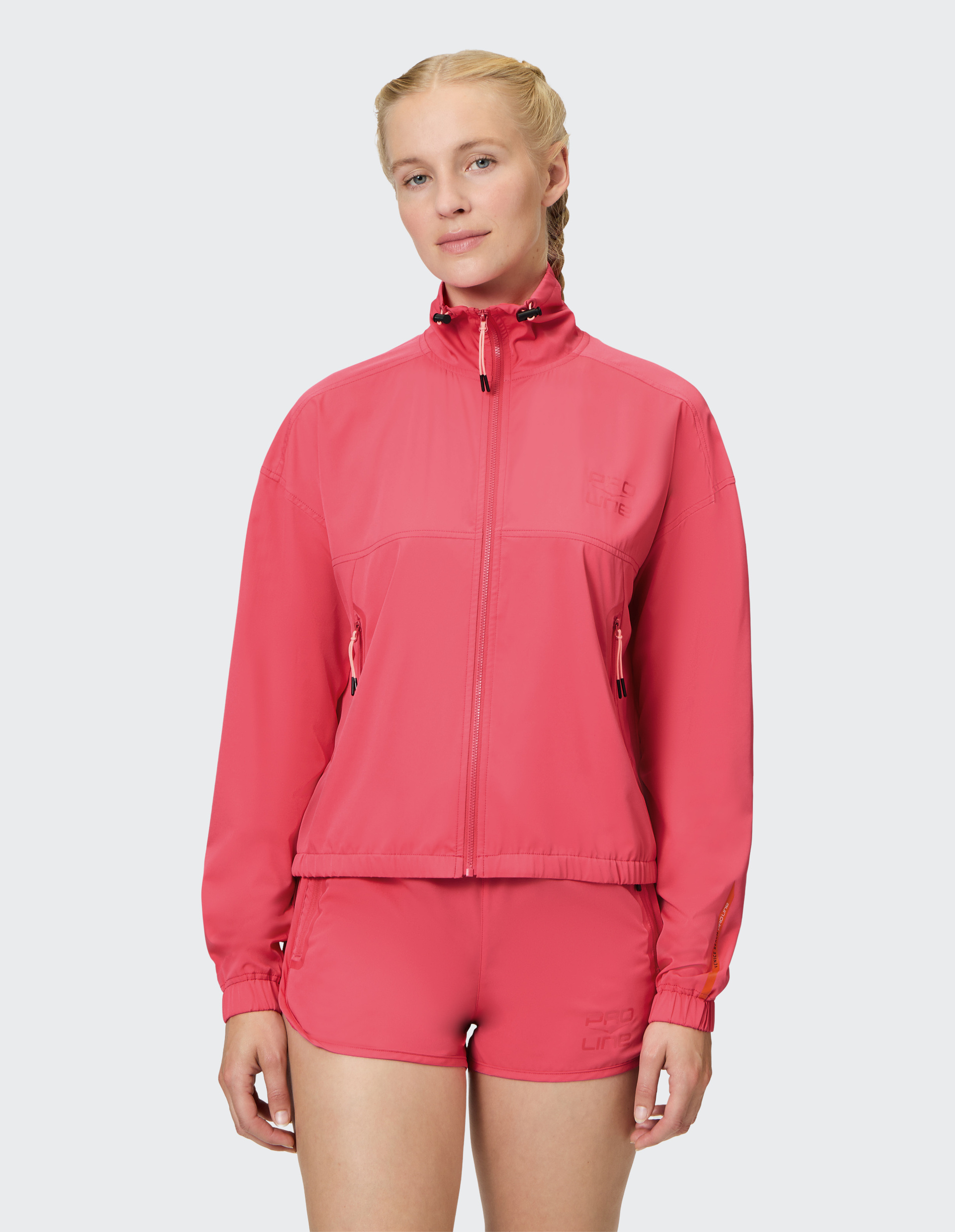 Frau mit blonden Zöpfen trägt eine rosa Sportjacke und passende Shorts, blickt entspannt in die Kamera.
