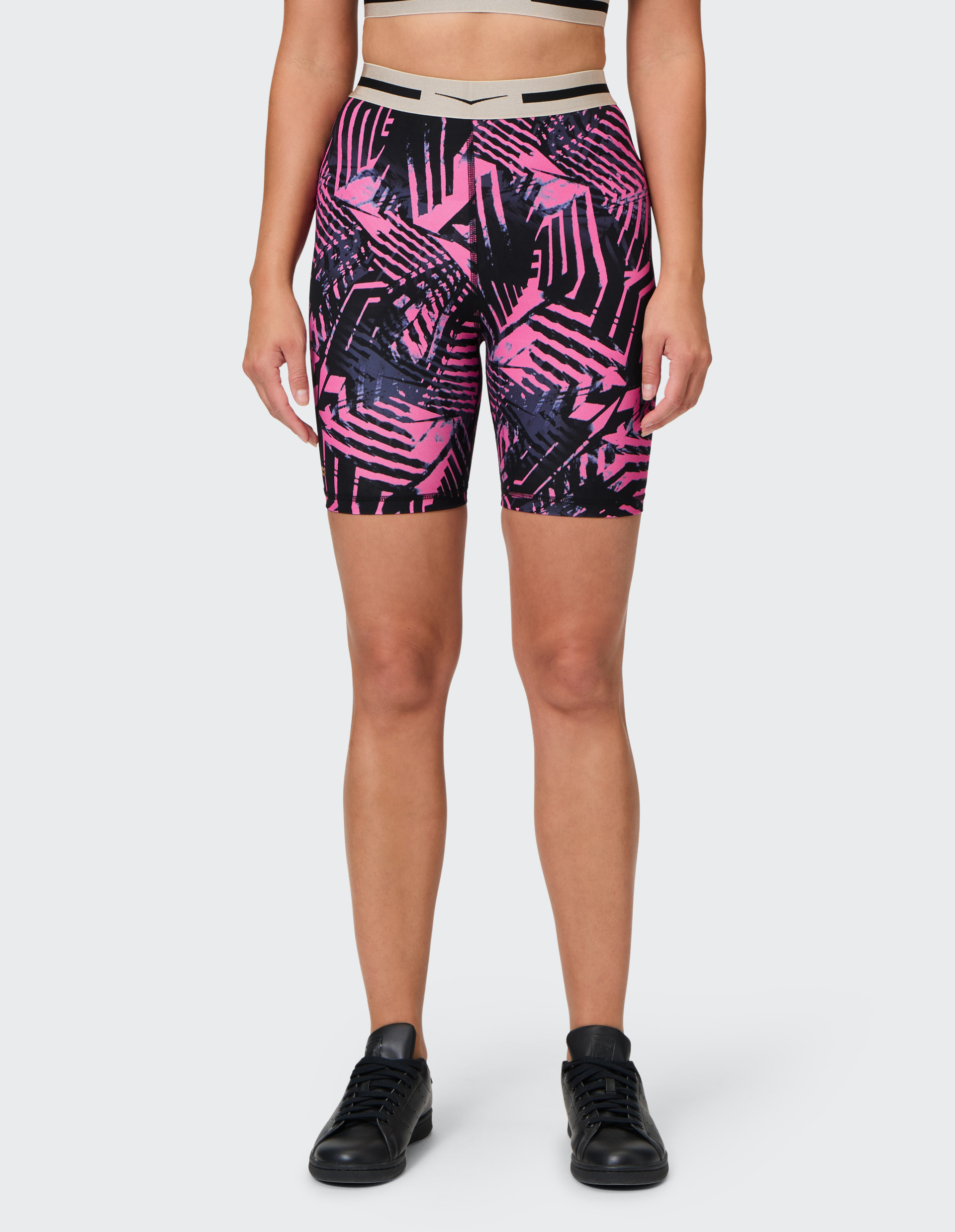 Frau trägt gemusterte, pink-schwarze Sportshorts und schwarze Sneaker, steht frontal mit neutralem Hintergrund.