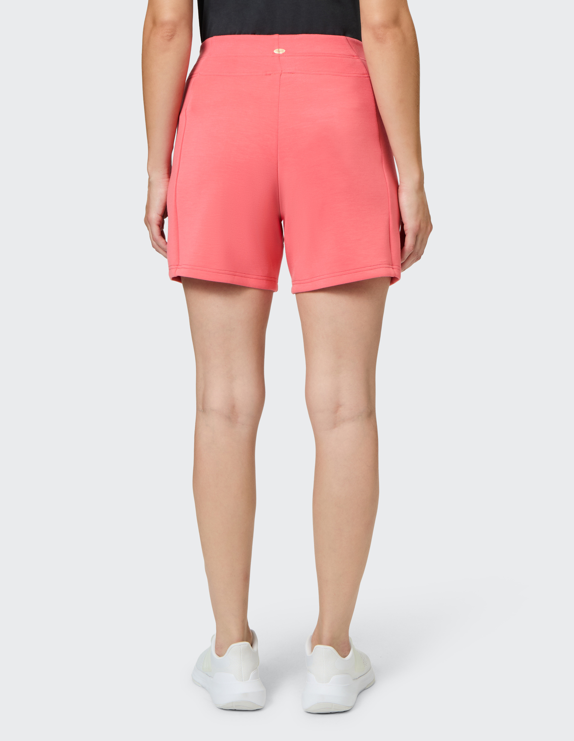 Rückansicht einer Person in einem schwarzen Oberteil, korallfarbenen Shorts und weißen Turnschuhen, stehend auf grauem Hintergrund.