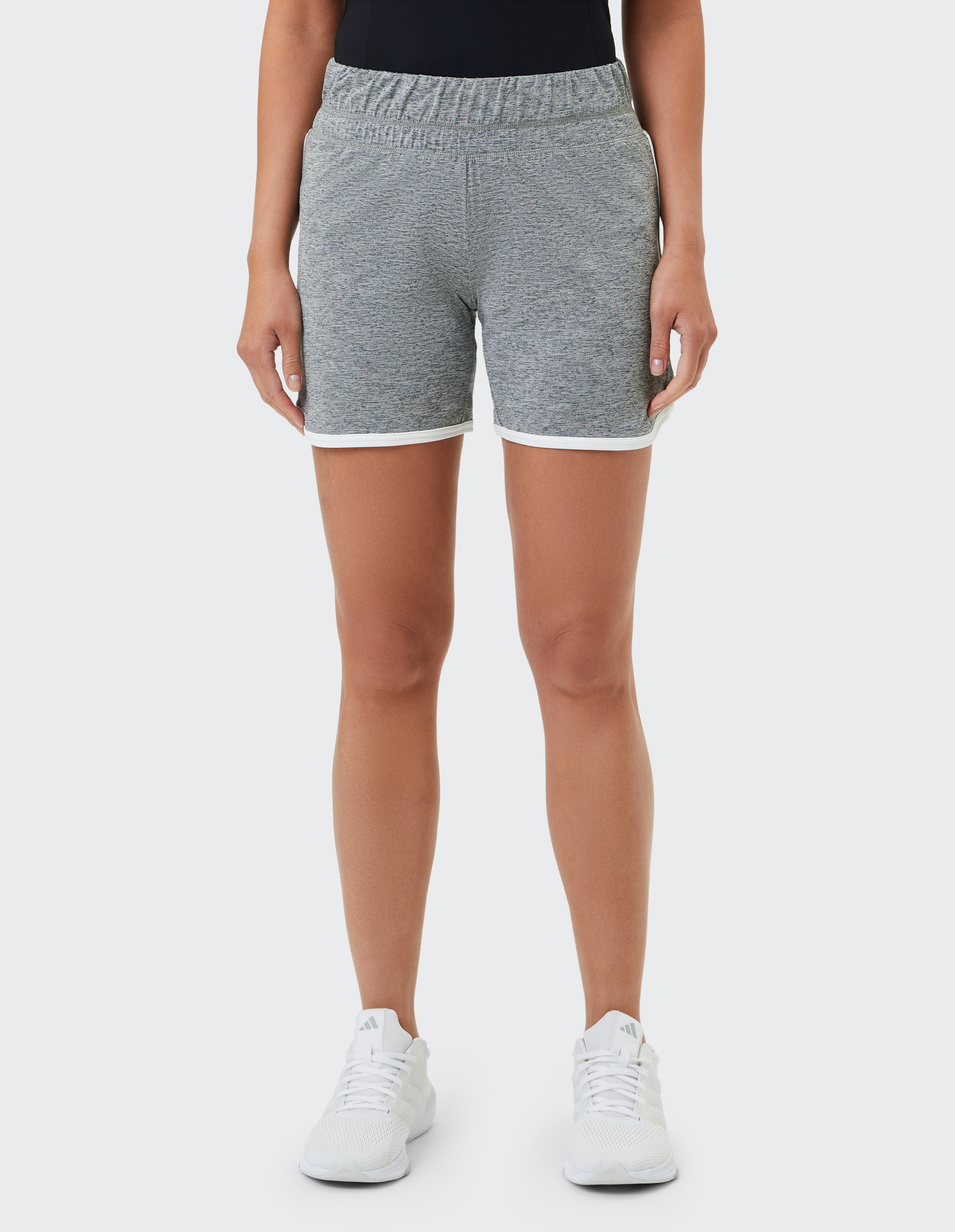 Frau trägt graue Shorts mit weißem Saum und weiße Sneaker, stehend vor einem neutralen Hintergrund.