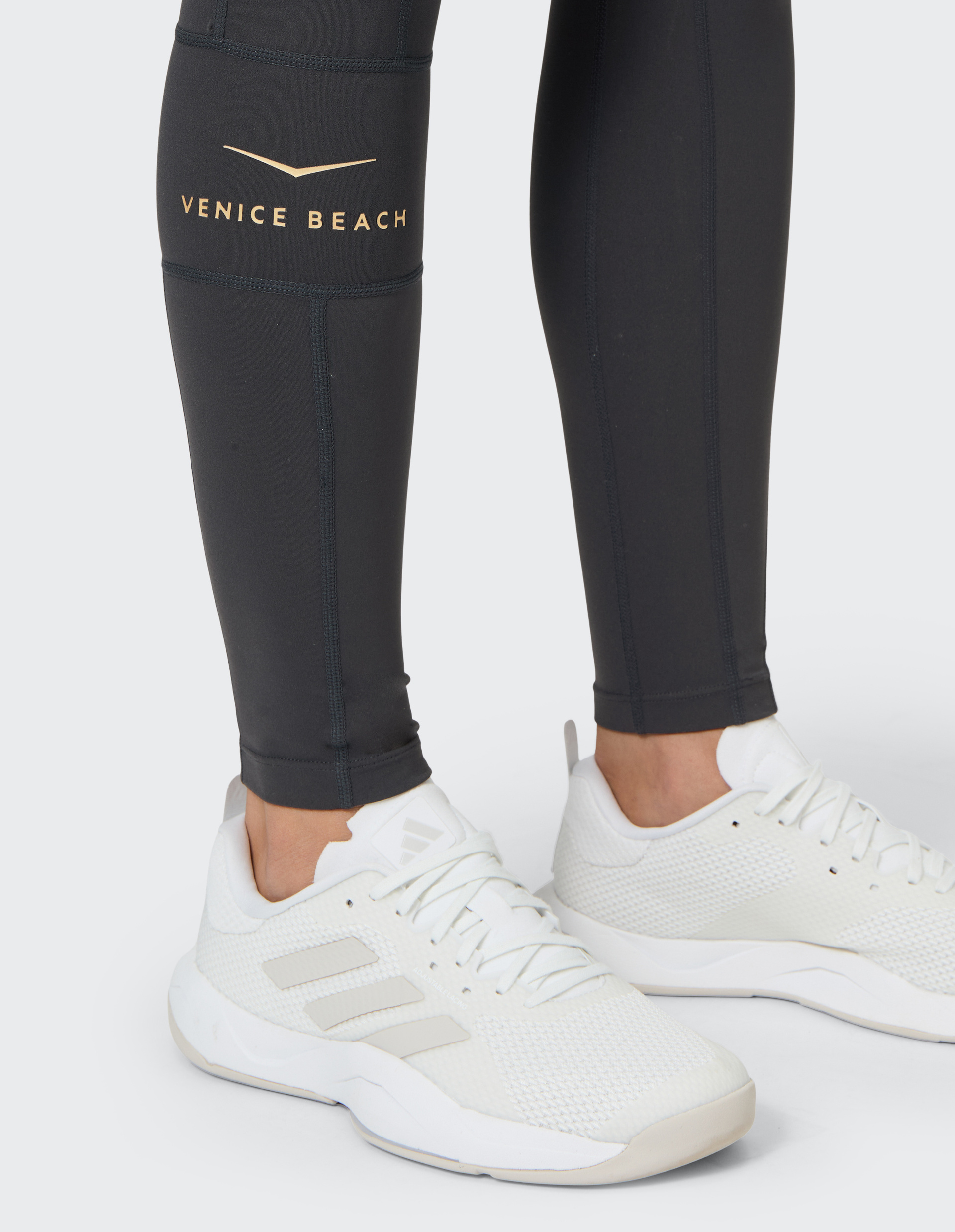 Weiße Sportschuhe und schwarze Leggings mit dem Schriftzug "Venice Beach" am Bein, Nahaufnahme der Beine auf hellgrauem Hintergrund.
