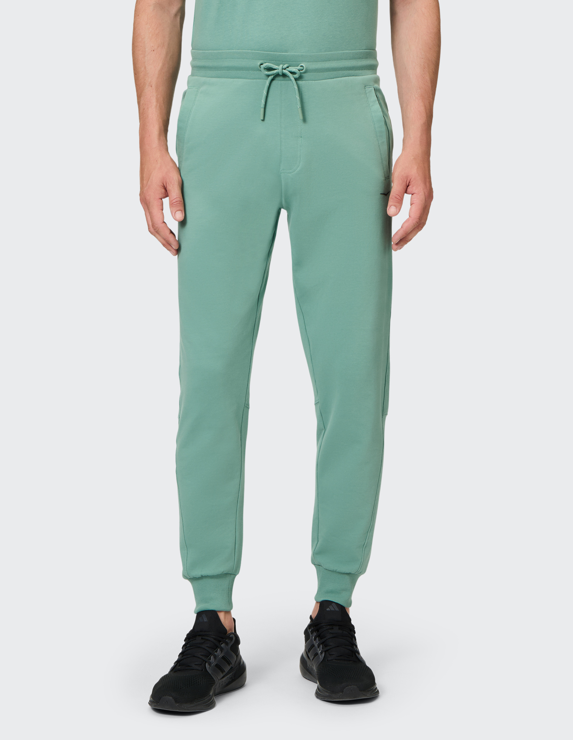 Mann trägt eine mintgrüne Jogginghose mit Kordelzug und Taschen, kombiniert mit schwarzen Sportschuhen vor weißem Hintergrund.