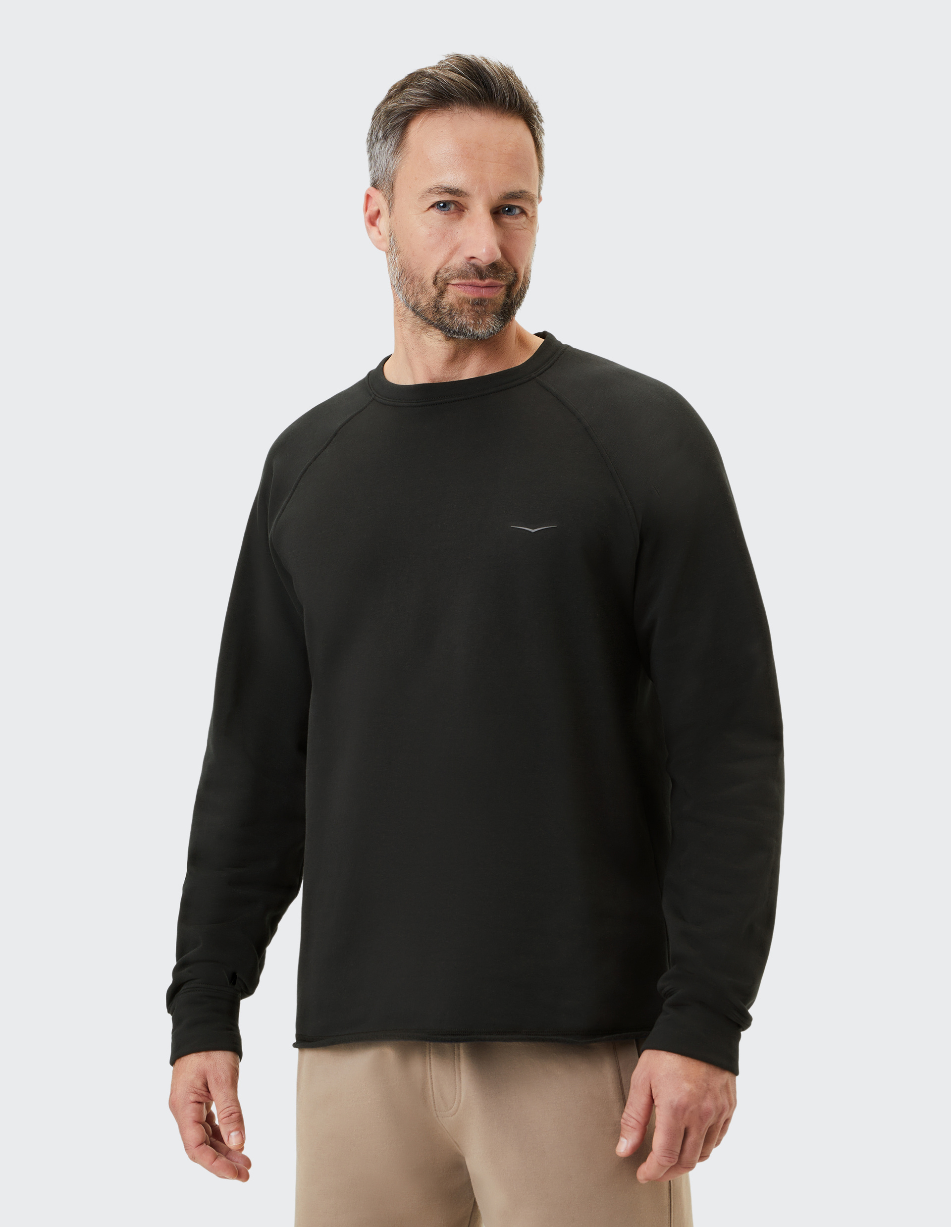 Ein Mann mit grauen Haaren trägt einen schwarzen Pullover und beige Hose vor einem hellgrauen Hintergrund.