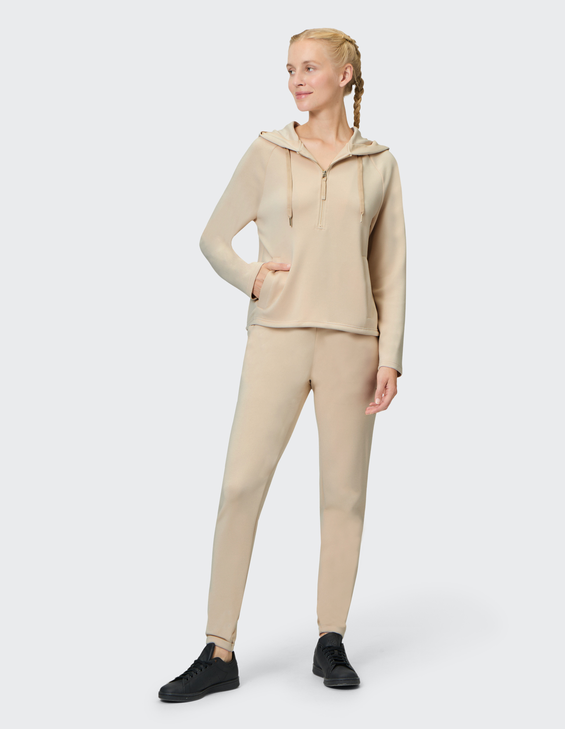 Frau in beigefarbenem Jogginganzug mit Kapuze und Reißverschluss, steht lächelnd seitlich, trägt schwarze Sneakers.