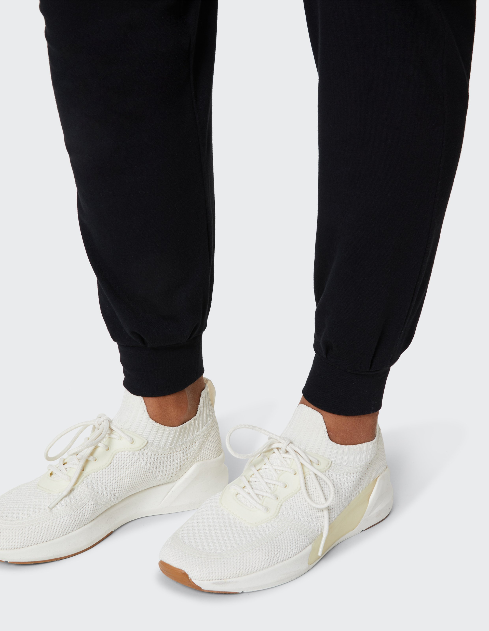 Ein Paar weiße Sneaker mit Strick-Obermaterial und Schnürsenkeln, getragen zu schwarzen Jogginghosen auf hellem Hintergrund.