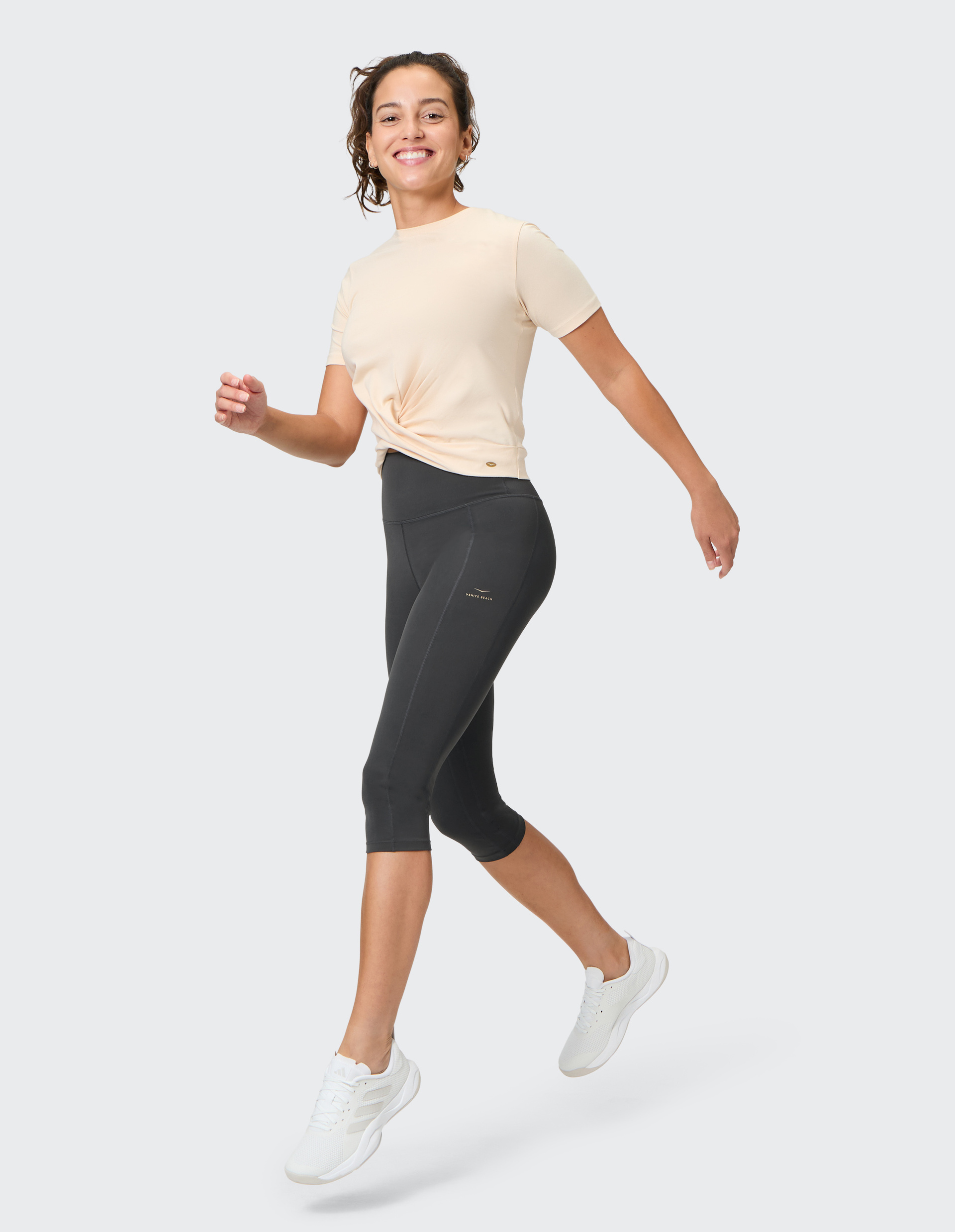 Frau in beige Shirt und schwarzen Leggings geht lächelnd, trägt weiße Sneaker, vor schlichtem grauem Hintergrund.