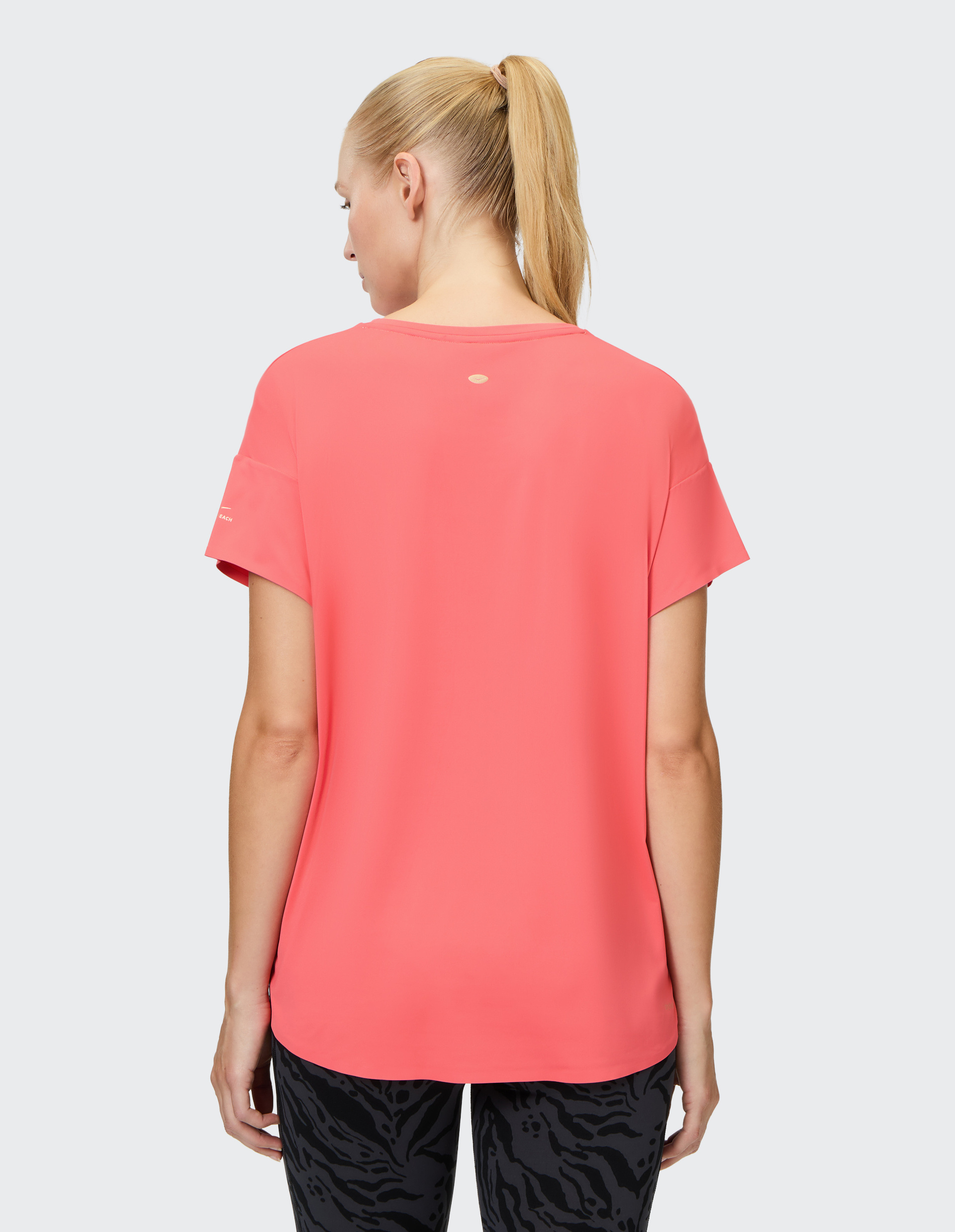 Frau mit blondem Pferdeschwanz trägt ein korallenrotes T-Shirt und schwarze gemusterte Leggings, von hinten fotografiert.