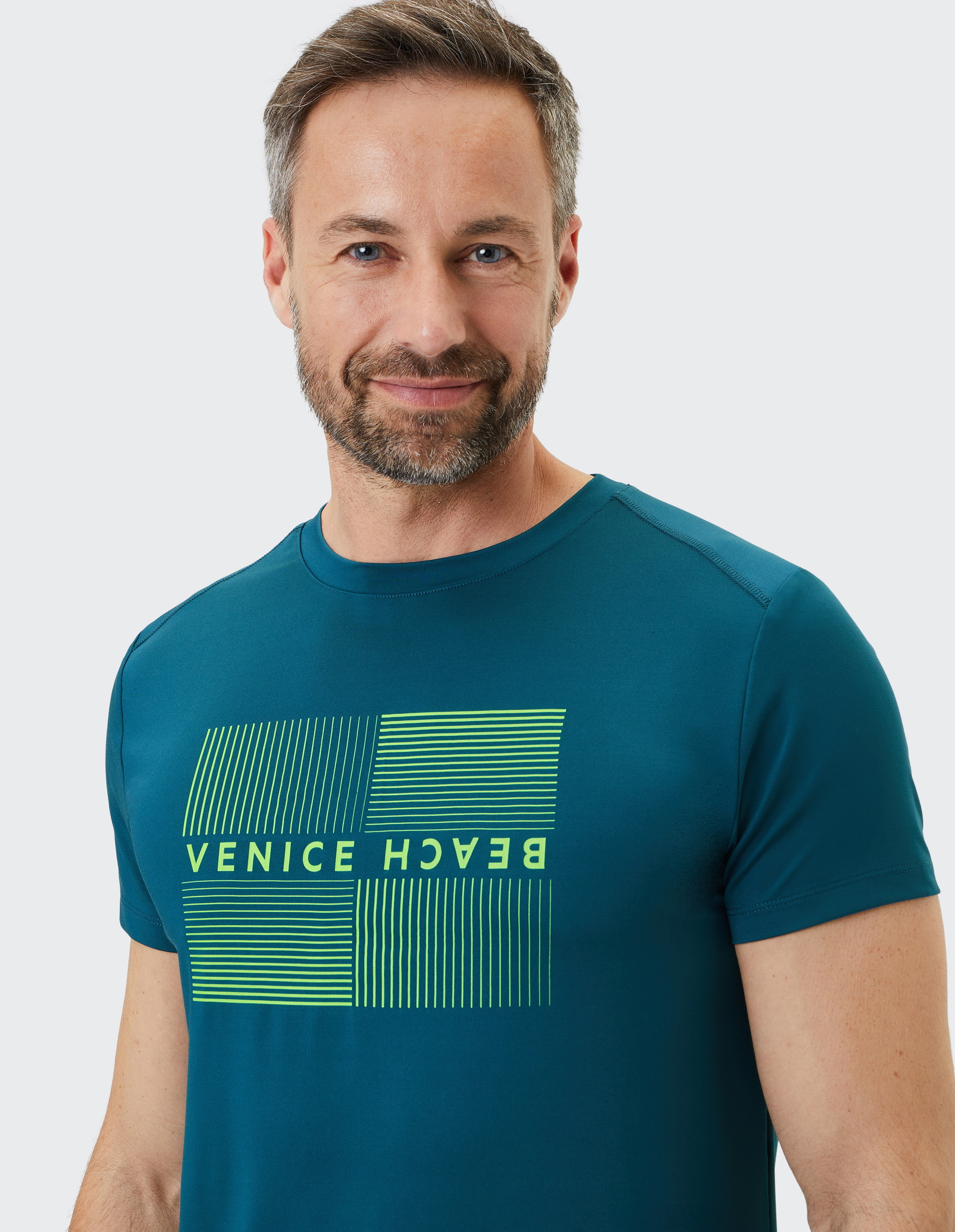 Mann mit kurzem Haar und Bart trägt ein blaues T-Shirt mit gelbem "Venice Beach"-Aufdruck, lächelnd vor hellem Hintergrund.