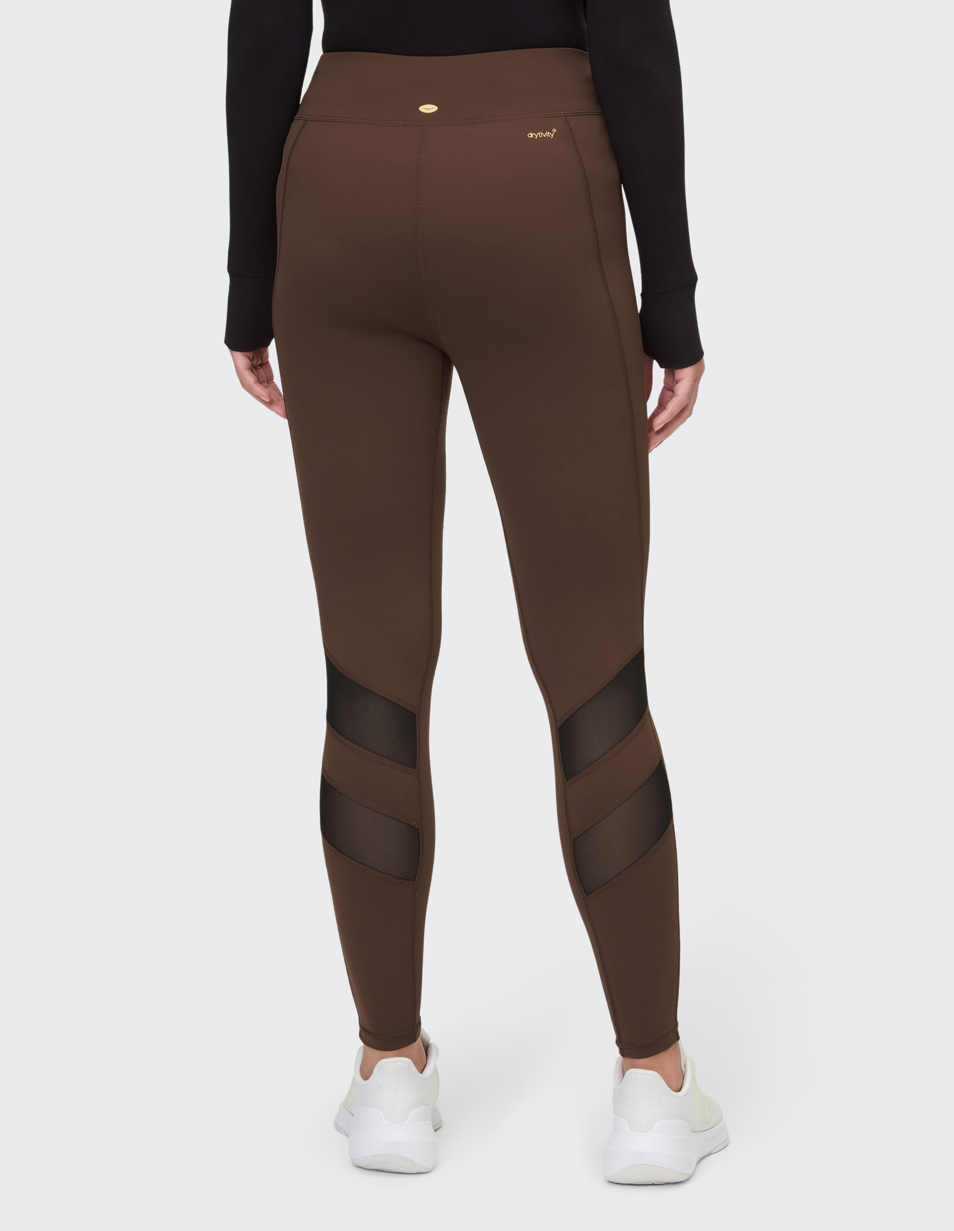 Rückansicht einer Person in brauner Leggings mit schwarzen Mesh-Einsätzen und weißen Sportschuhen auf hellem Hintergrund.