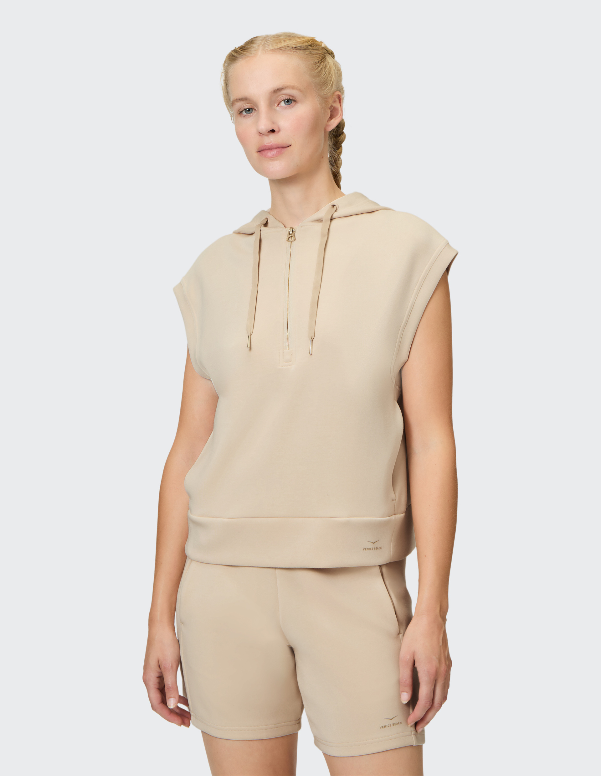 Frau in beigefarbenem ärmellosen Kapuzenpullover mit halbem Reißverschluss und passenden Shorts, geflochtenes Haar, neutraler Hintergrund.