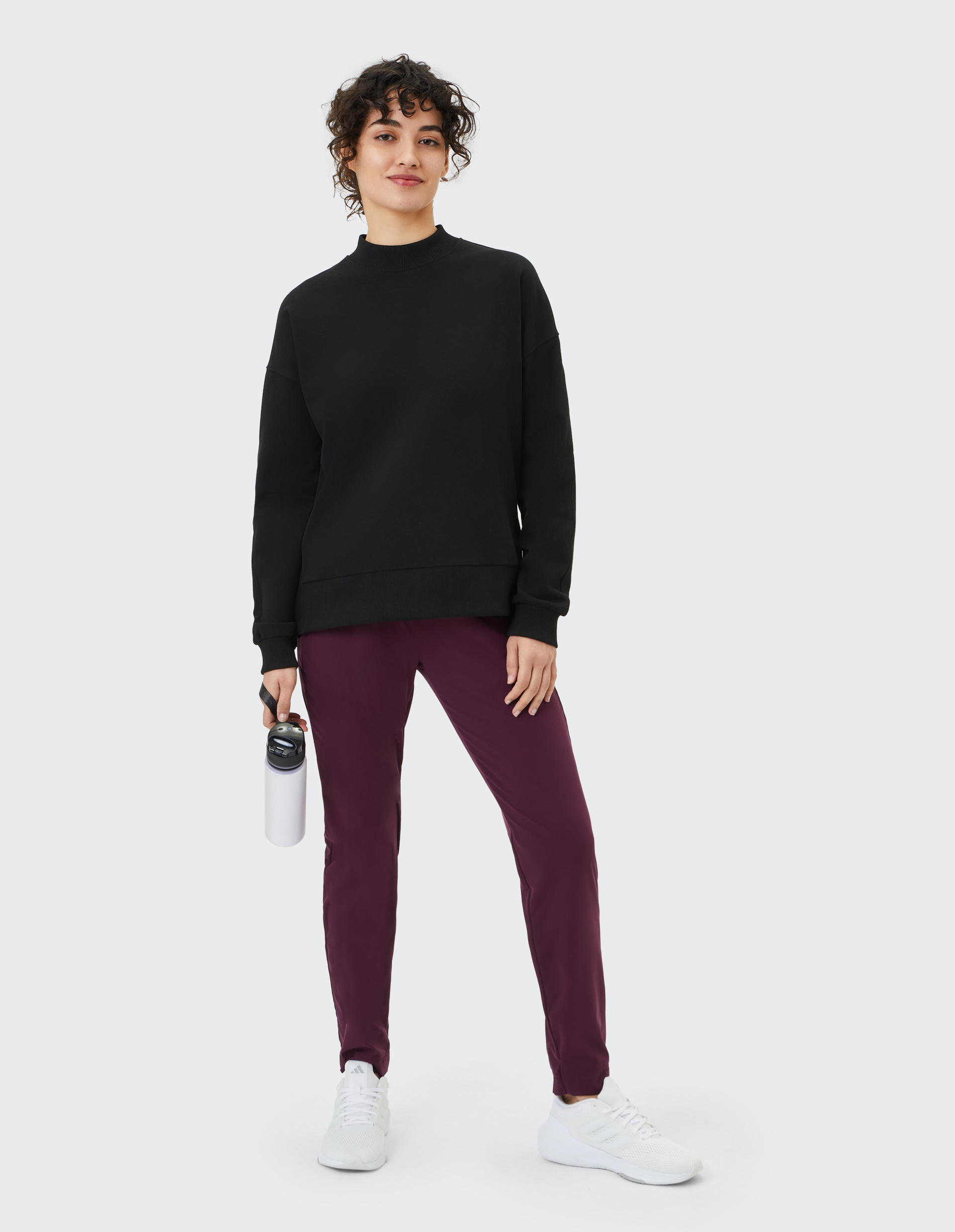 Frau in schwarzem Pullover und burgunderroter Hose hält eine weiße Wasserflasche, steht aufrecht und lächelt, vor grauem Hintergrund.