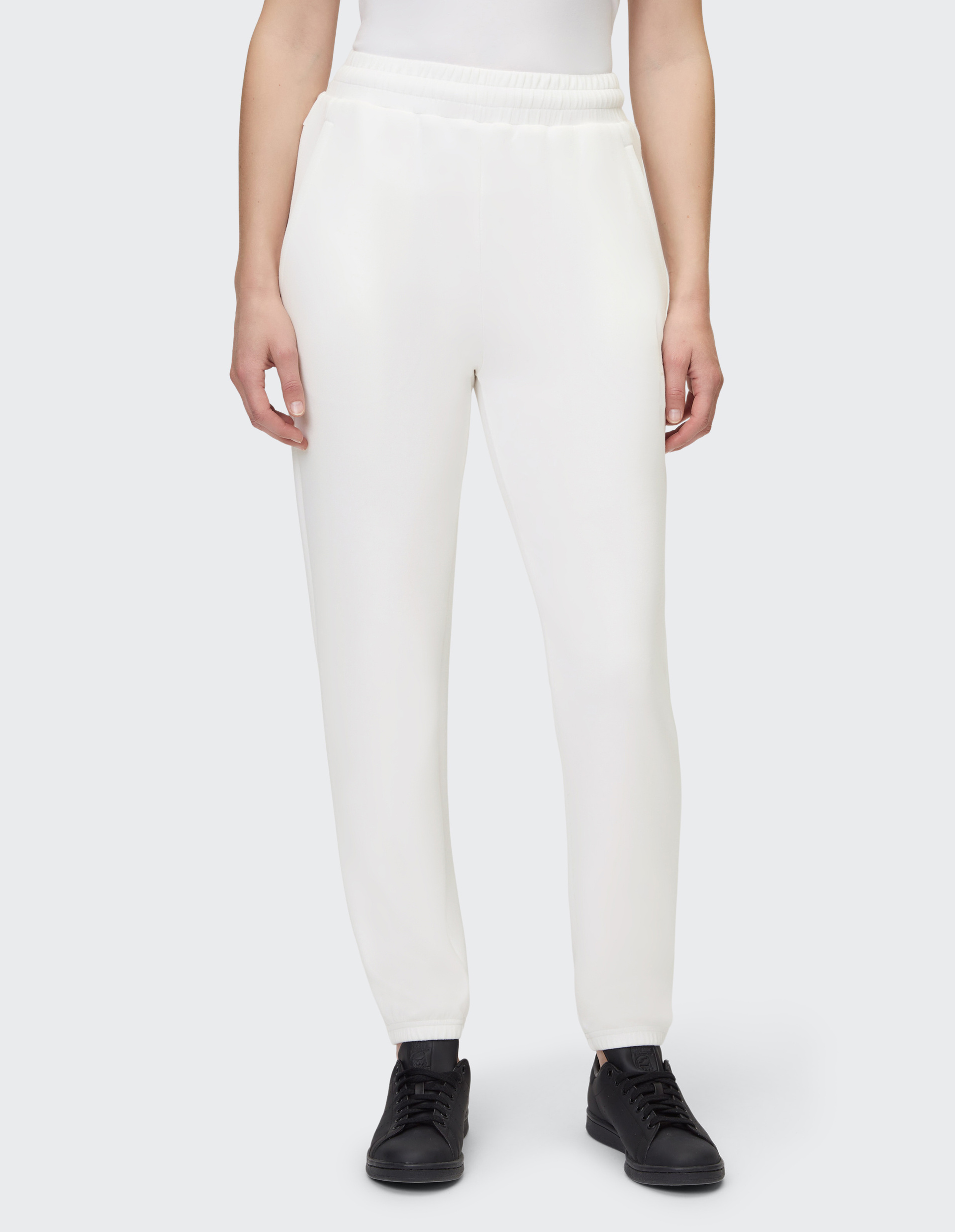 Eine Person trägt eine weiße Jogginghose mit elastischem Bund und schwarzen Sneakern auf grauem Hintergrund, Hüfte bis Füße sichtbar.