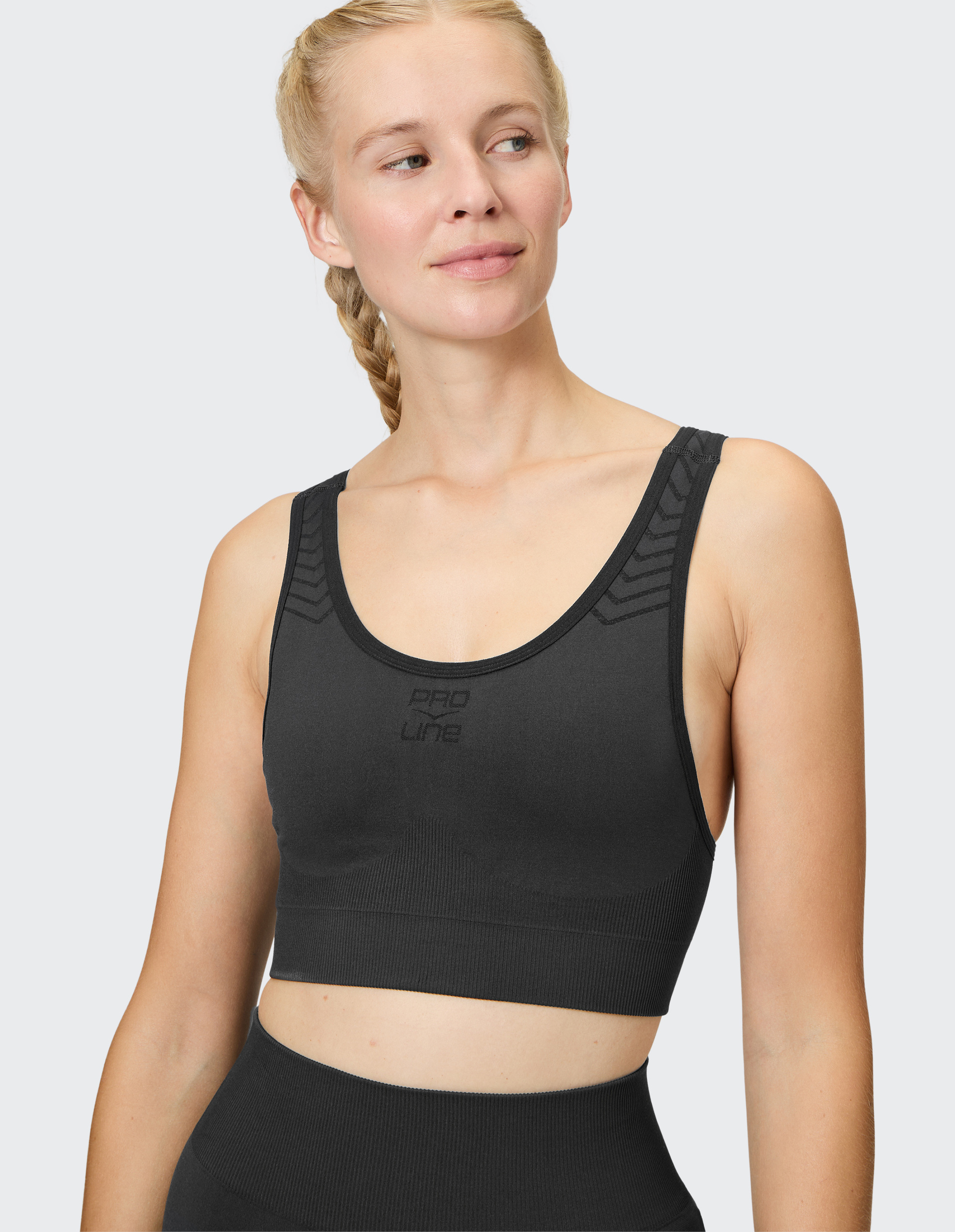 Frau mit geflochtenem Zopf trägt schwarzes Sporttop und blickt lächelnd nach rechts, vor grauem Hintergrund.