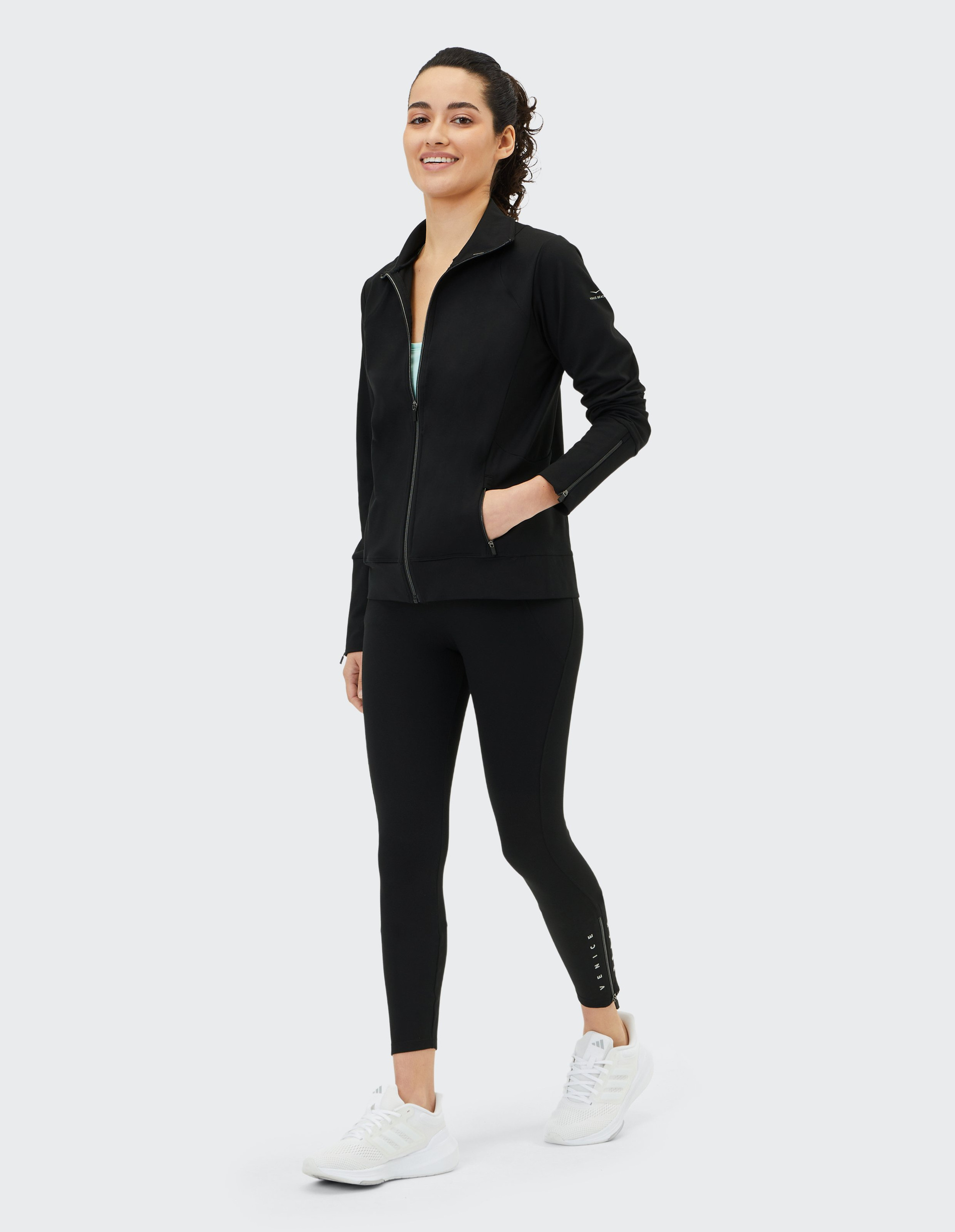 Junge Frau in schwarzem Sportanzug und weißen Sneakers lächelt und geht vor hellem Hintergrund.