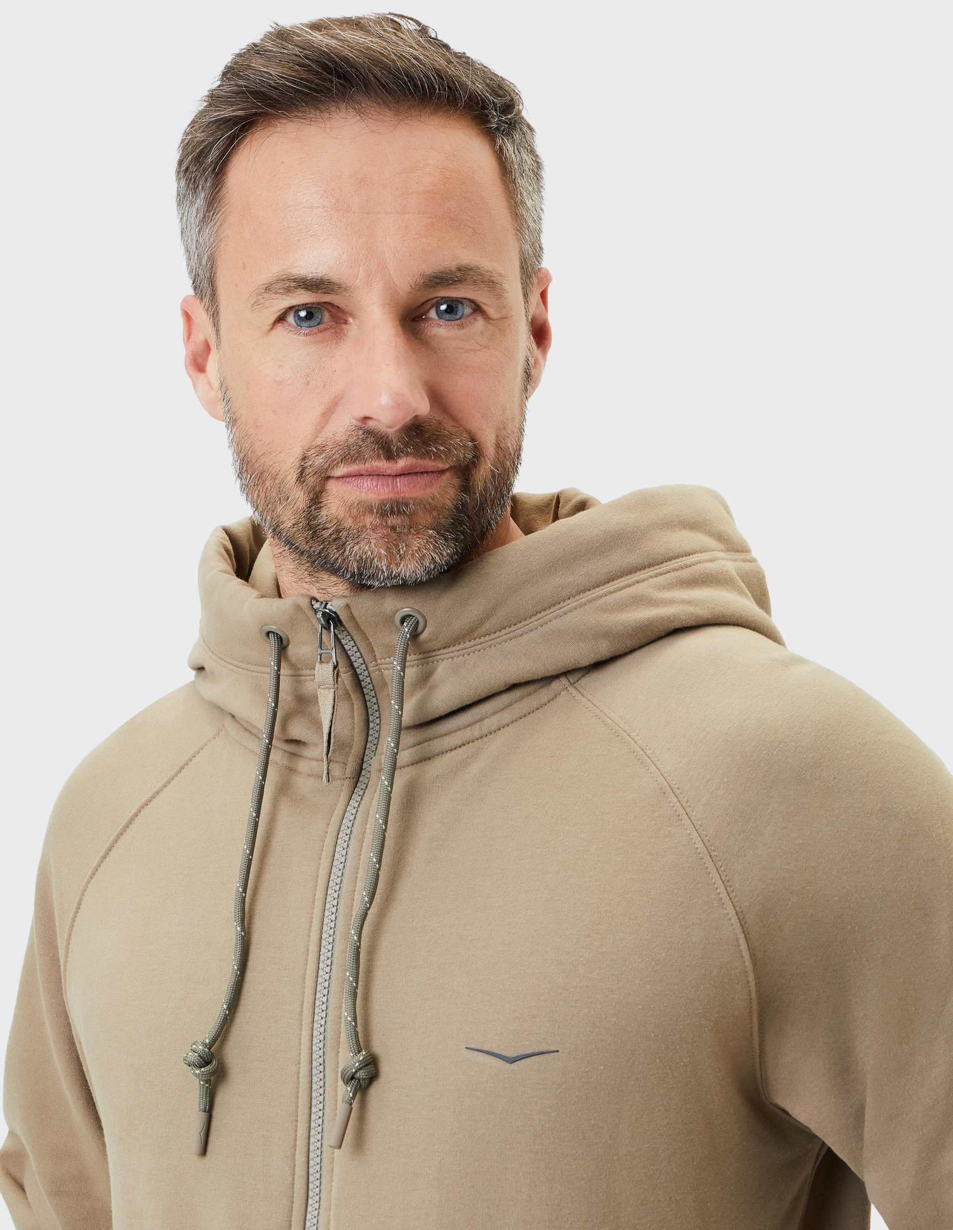 Mann mit kurzem Bart trägt beige Kapuzenpullover mit Kordeln. Er schaut lächelnd in die Kamera, Hintergrund weiß.
