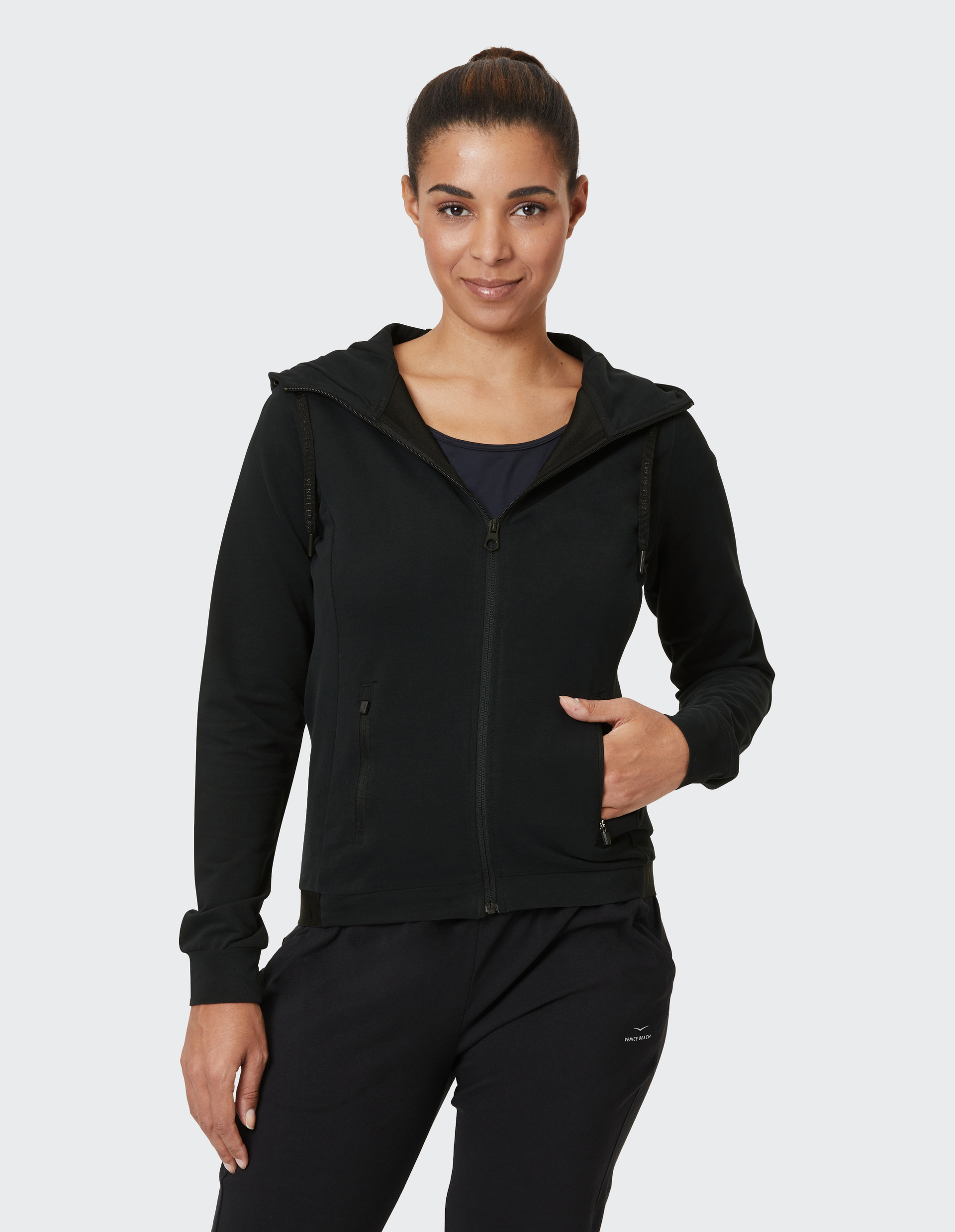 Eine Person trägt eine schwarze Kapuzenjacke und Hose, die entspannt dasteht und freundlich lächelt vor einem hellen Hintergrund.