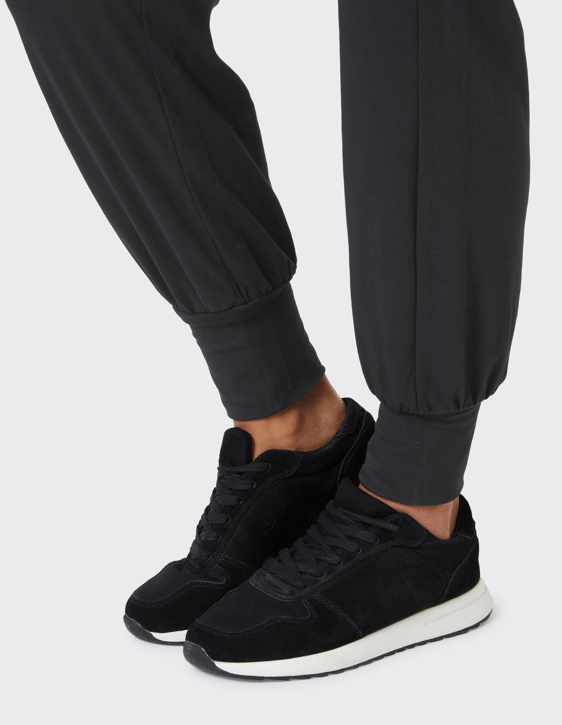 Schwarze Sneaker mit weißen Sohlen und schwarzer Jogginghose mit Bündchen, die leicht Falten wirft, auf hellem Hintergrund.