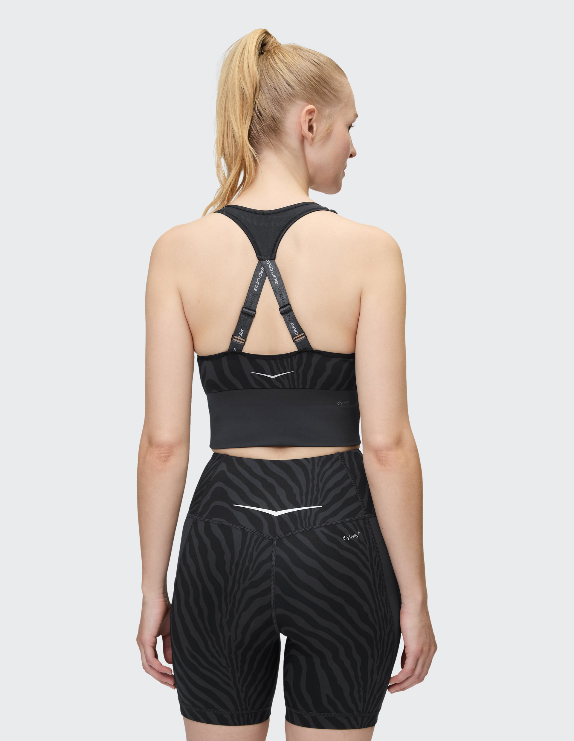 Frau mit blondem Pferdeschwanz trägt einen schwarzen Sport-BH und Shorts mit Zebra-Muster, Rückenansicht auf weißem Hintergrund.
