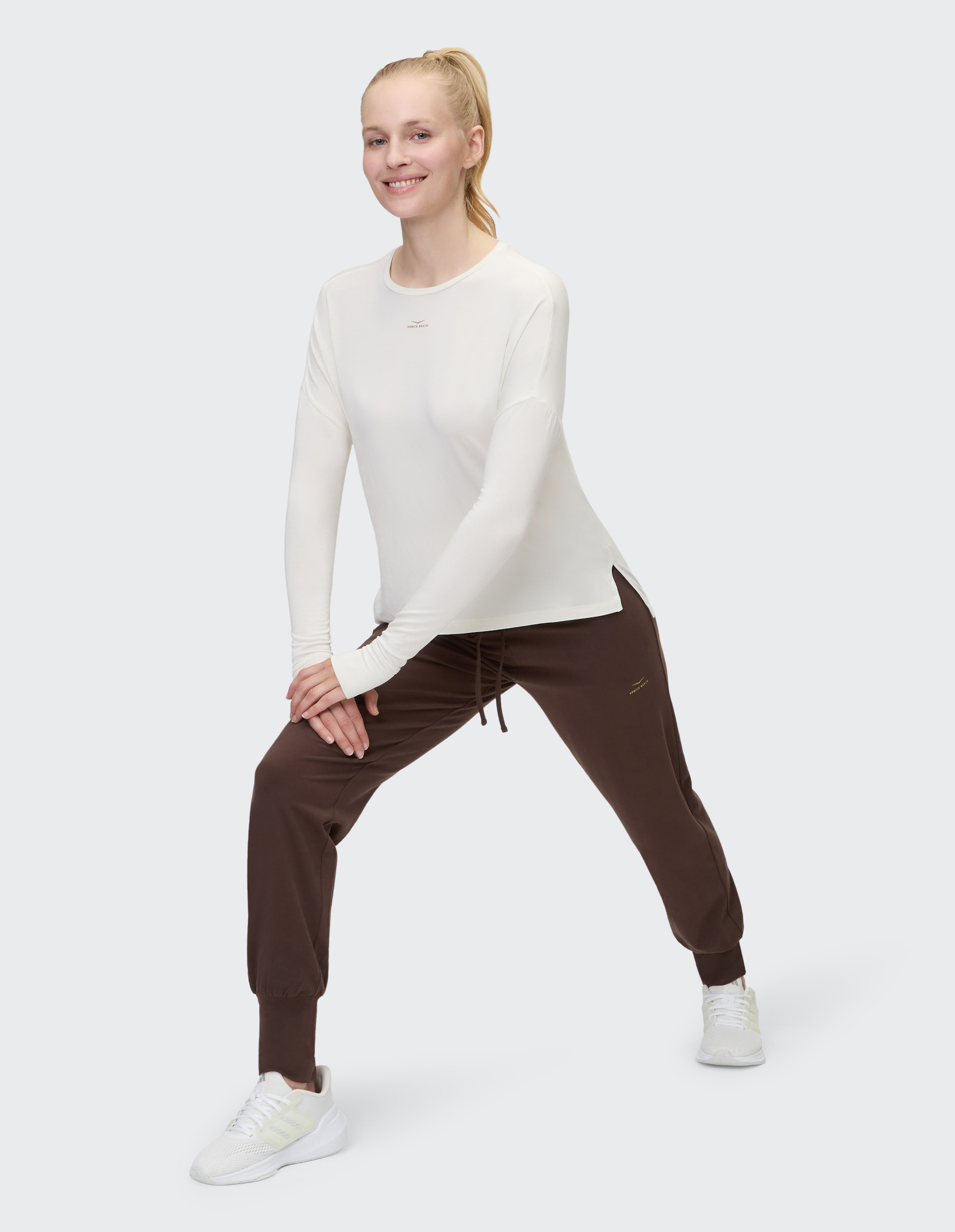 Eine Person trägt ein weißes Langarmshirt und braune Jogginghosen, dazu weiße Turnschuhe, stehend vor hellem Hintergrund.