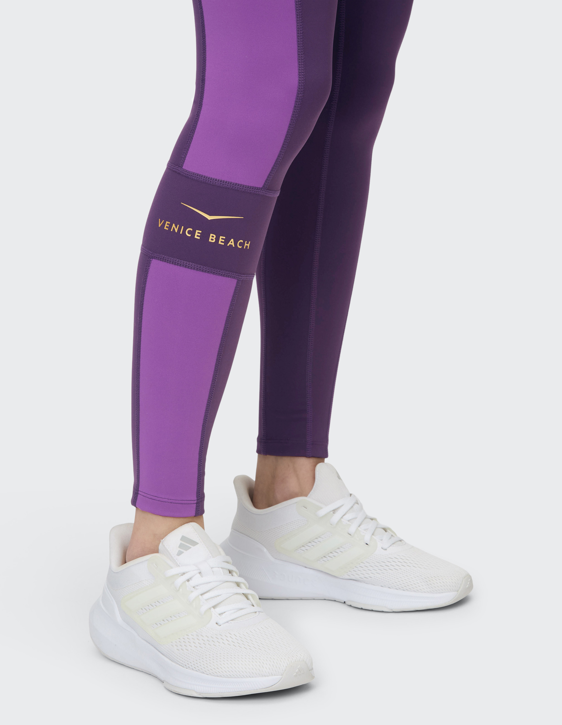 Person trägt lila Leggings mit Venice Beach-Logo und weiße Sneakers auf hellem Hintergrund.