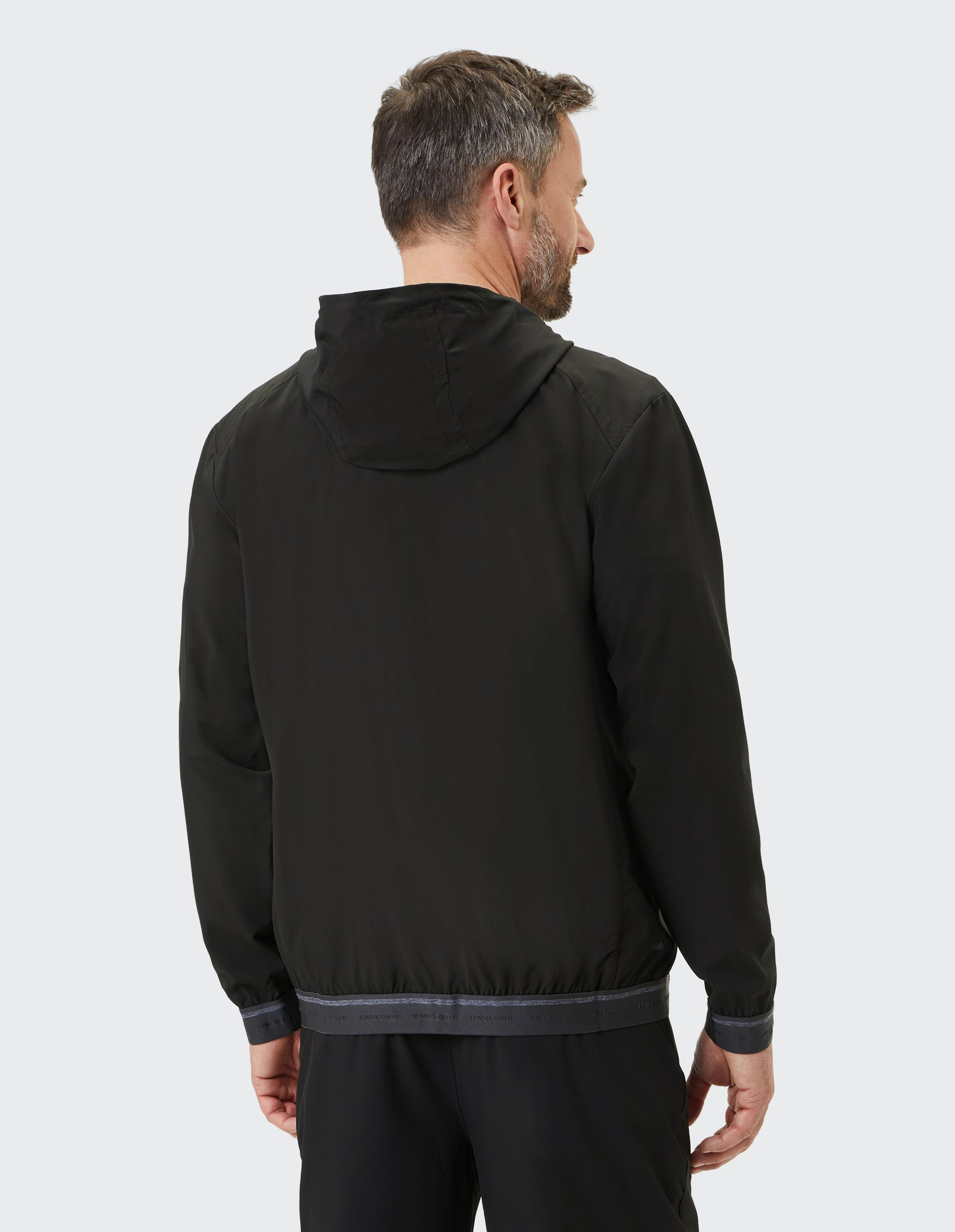 Ein Mann in schwarzer Jacke mit Kapuze steht rückwärts vor grauem Hintergrund und blickt nach rechts.