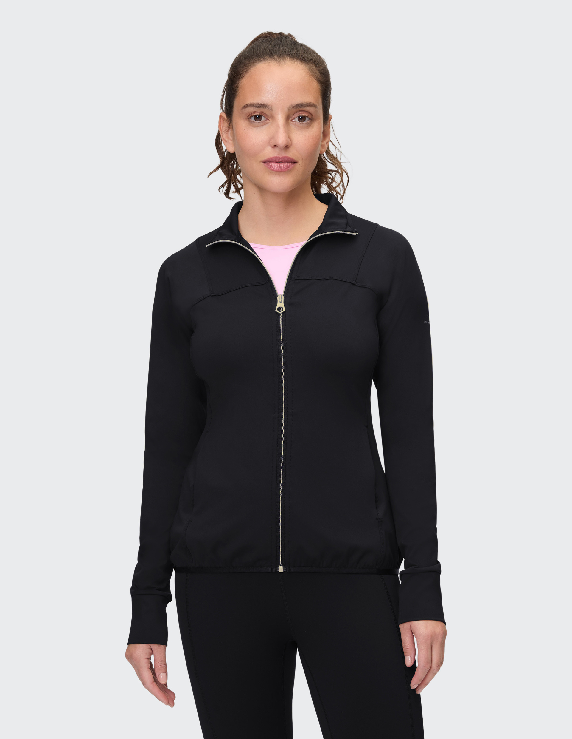 Frau mit lockigem Haar trägt eine schwarze Jacke mit Reißverschluss und rosa T-Shirt, steht frontal vor grauem Hintergrund.