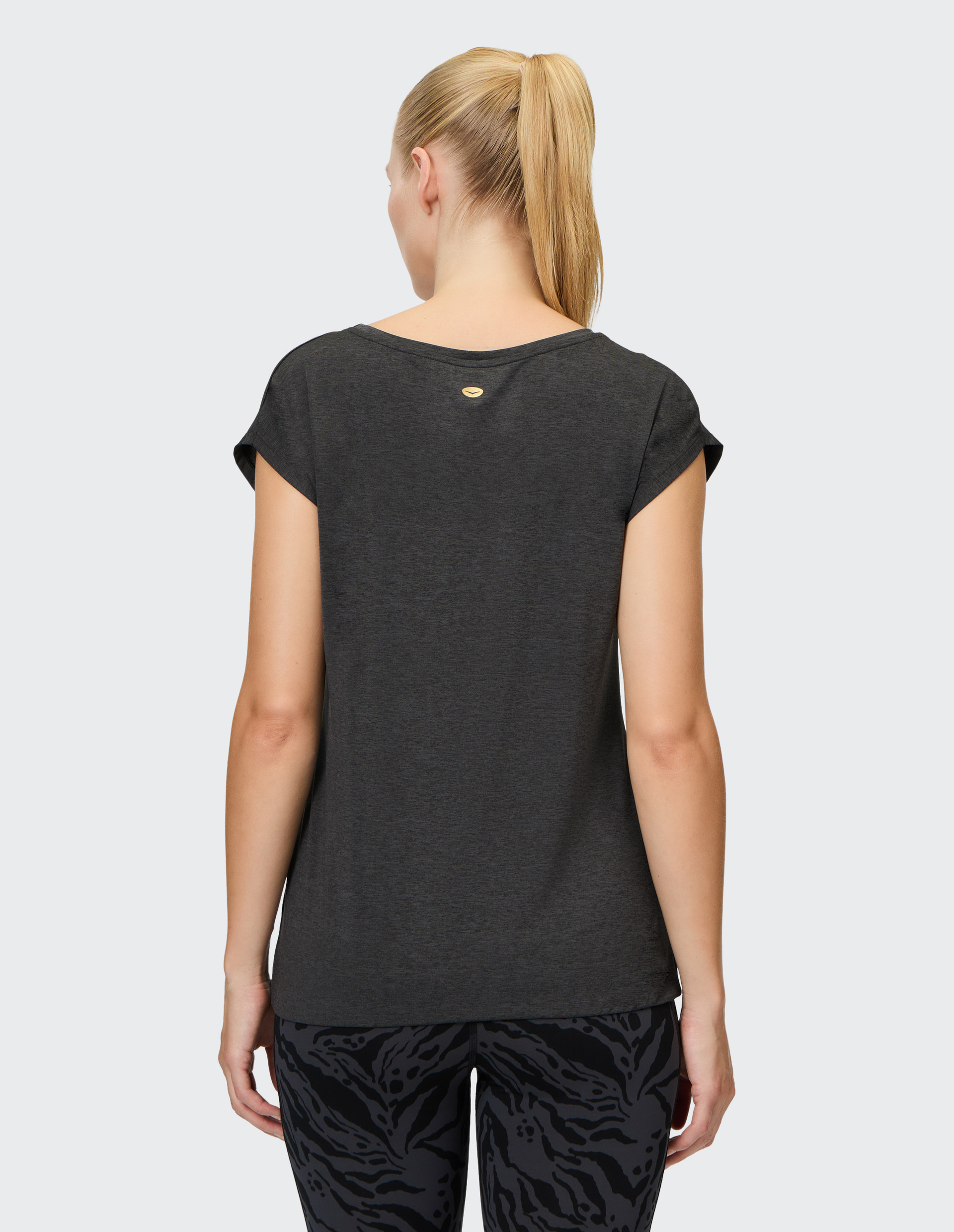 Eine Frau mit blondem Pferdeschwanz trägt ein dunkelgraues Shirt und gemusterte Leggings, von hinten fotografiert auf grauem Hintergrund.