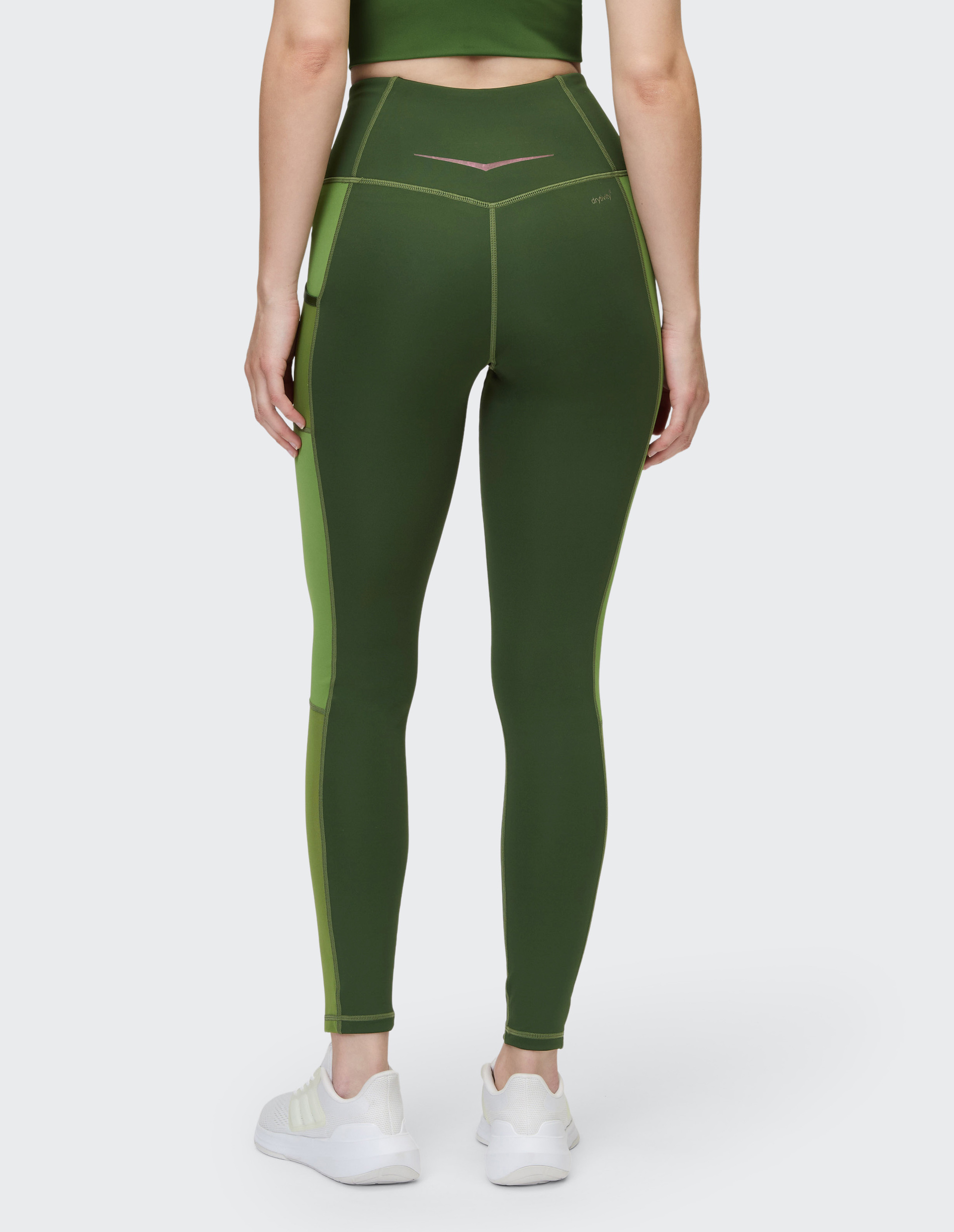 Eine Frau in grüner Sportbekleidung von hinten, trägt High-Waist-Leggings und weiße Turnschuhe, stehend auf hellem Hintergrund.