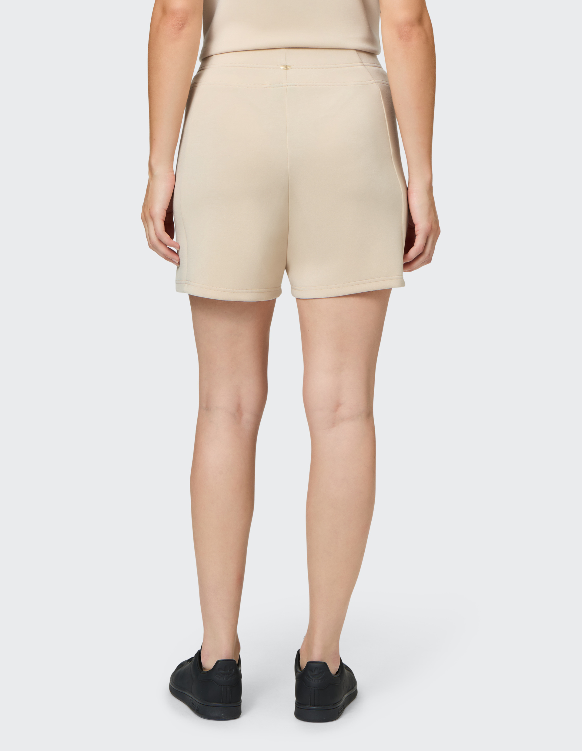 Rückansicht einer Person in beigefarbenem Oberteil und kurzen Shorts, schwarze Sneakers, vor hellem Hintergrund stehend.