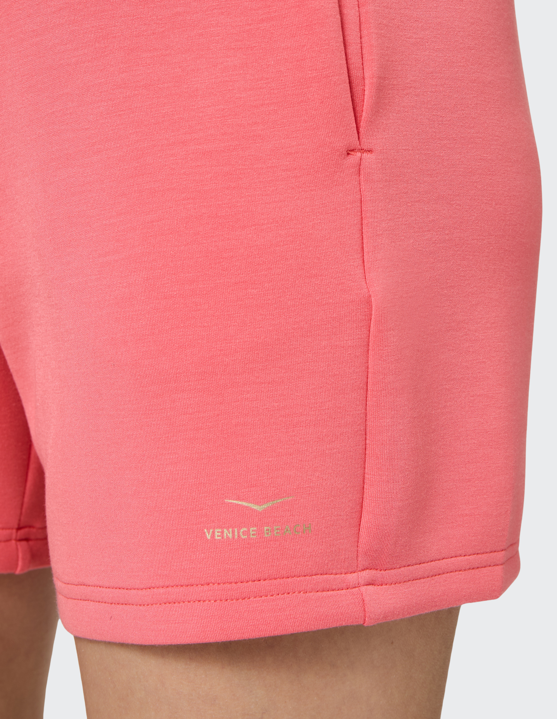 Nahaufnahme einer korallfarbenen Shorts mit dezenter Seitentasche und kleinem Venice Beach-Logo am unteren Saum auf beigefarbenem Hintergrund.