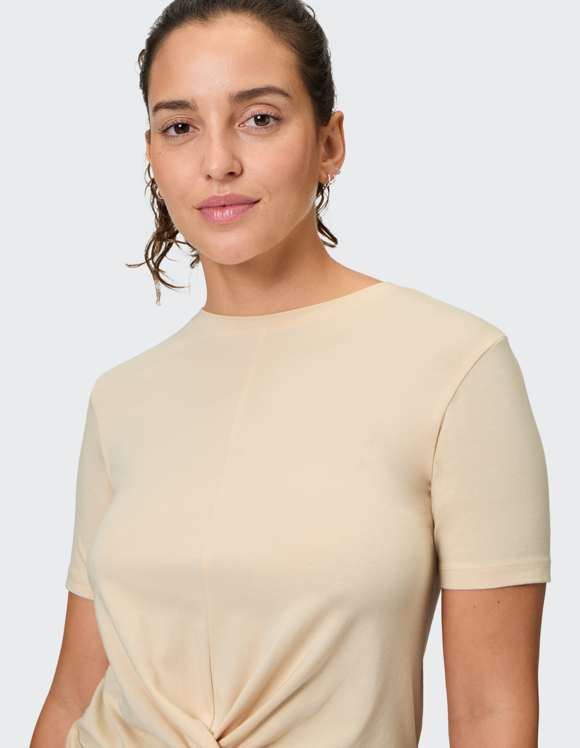 Frau mit lockigem Haar in beigefarbenem T-Shirt lächelt sanft vor grauem Hintergrund, trägt dezente Ohrringe.