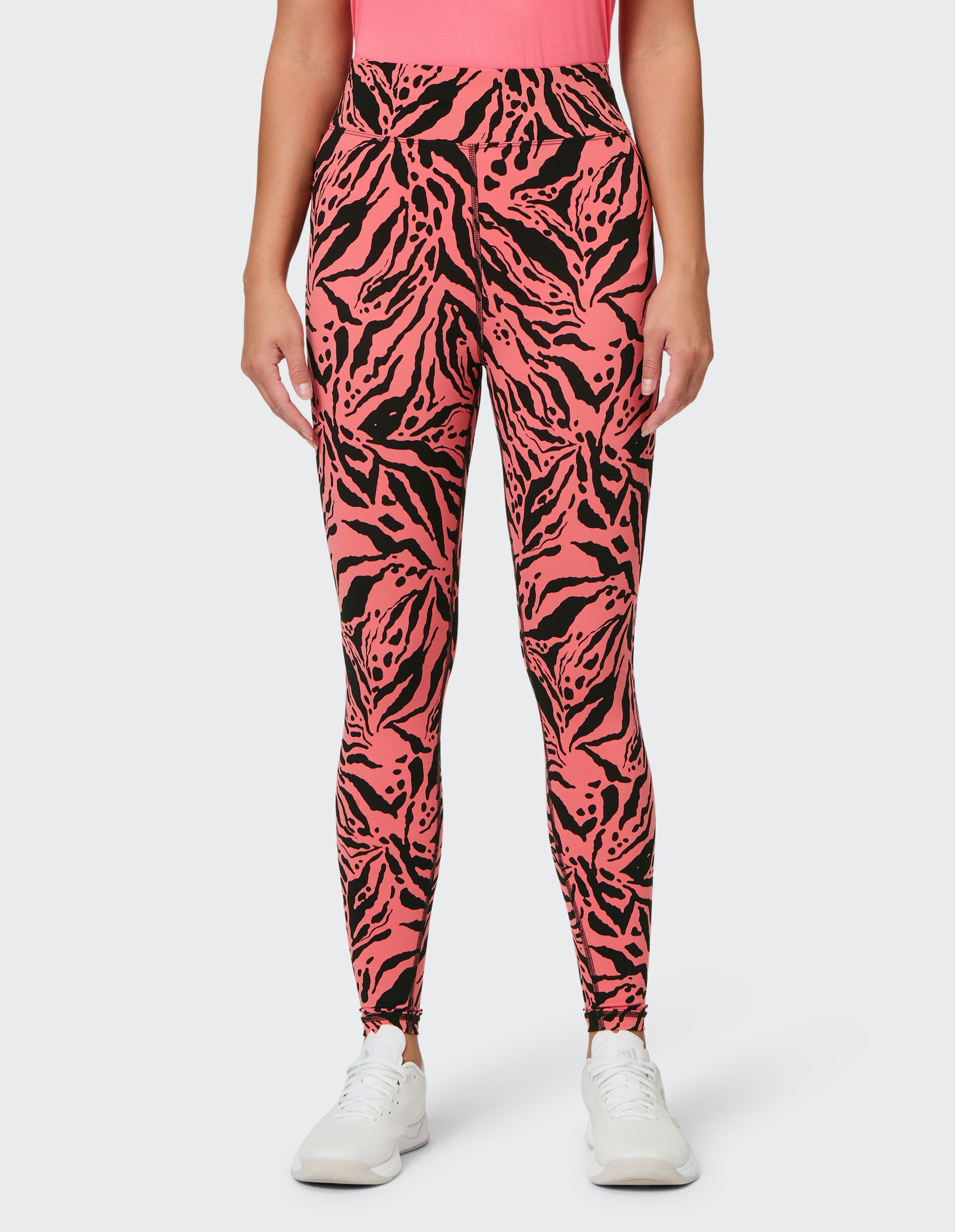 Frau trägt pinke Leggings mit schwarzem, abstraktem Muster und ein pinkes Oberteil, dazu weiße Sportschuhe, vor grauem Hintergrund.