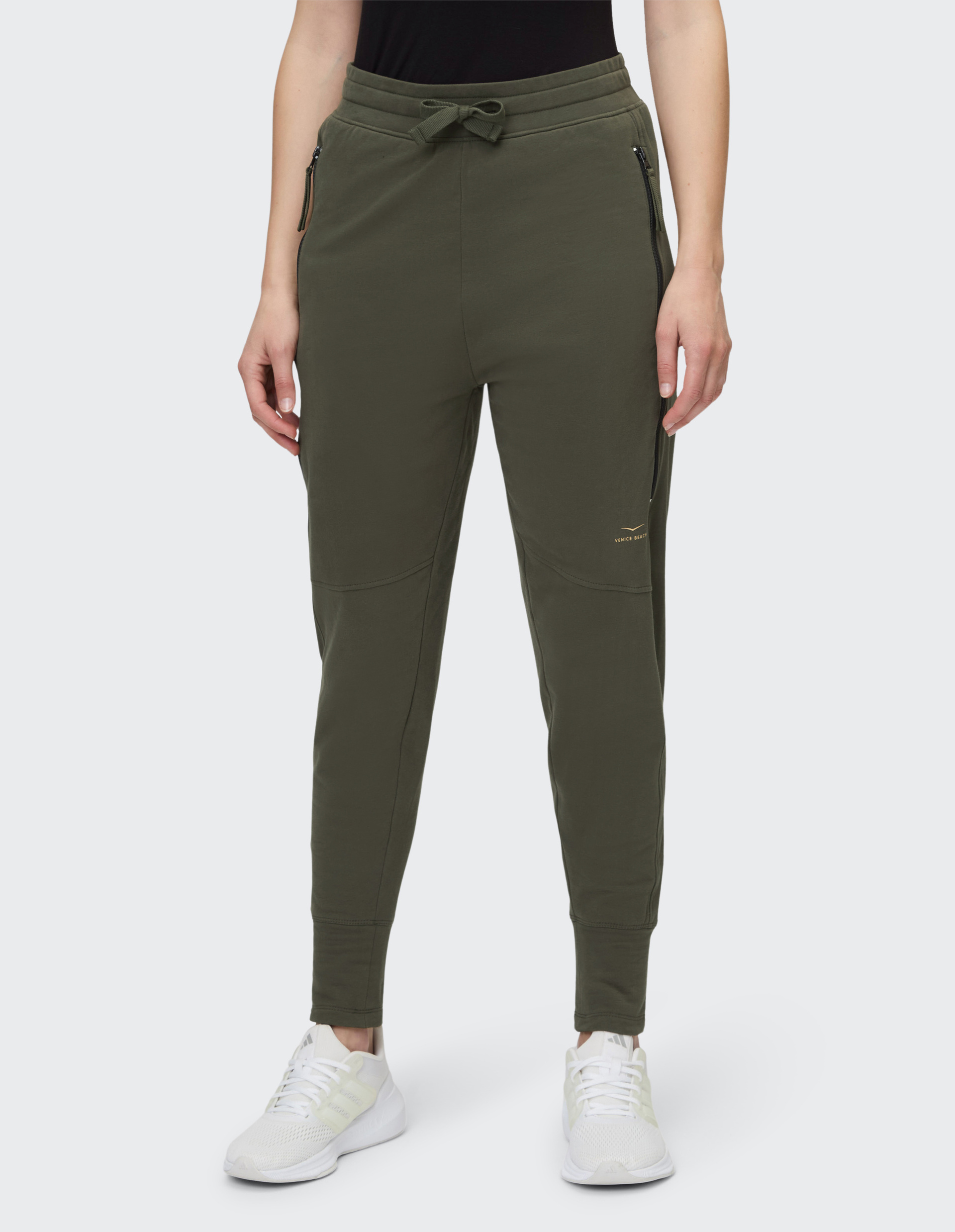 Person trägt olivgrüne Jogginghose mit Kordelzug, seitlichen Reißverschlusstaschen, kombiniert mit weißen Turnschuhen.