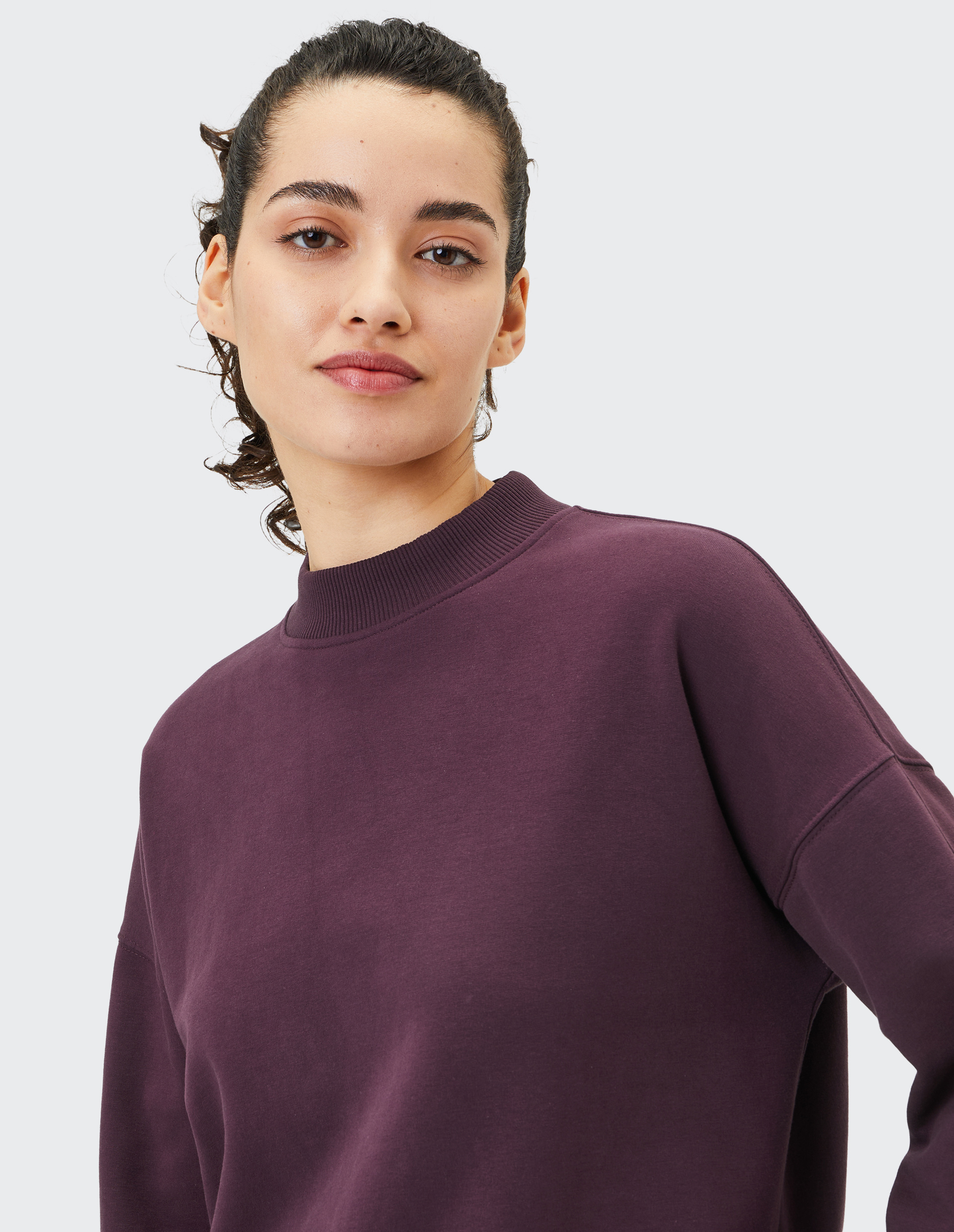 Frau mit zurückgebundenem Haar trägt einen dunkelvioletten Pullover und blickt freundlich lächelnd mit hellem Hintergrund in die Kamera.