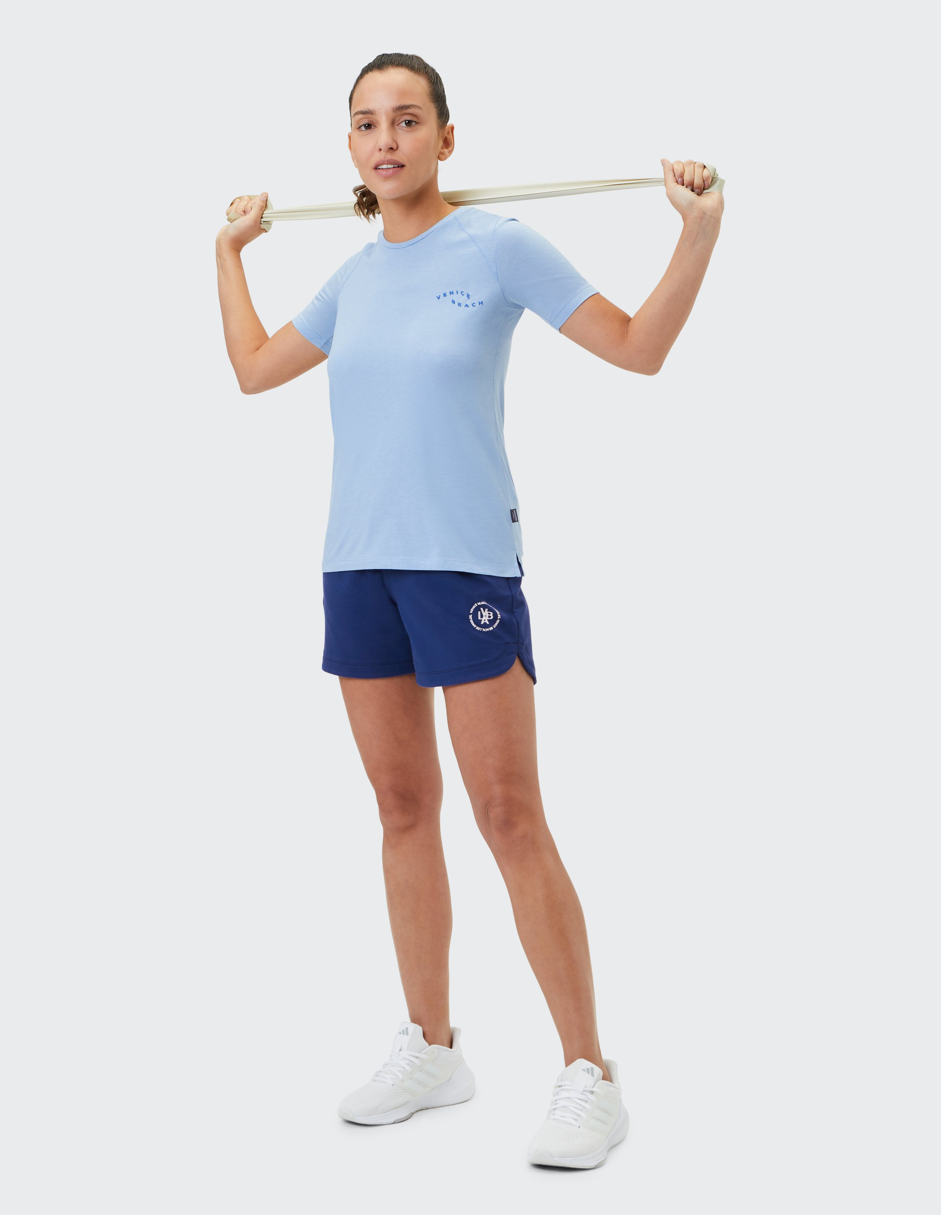 Frau in hellblauem T-Shirt und blauen Shorts hält ein elastisches Band über den Schultern, bereit für ein Workout.