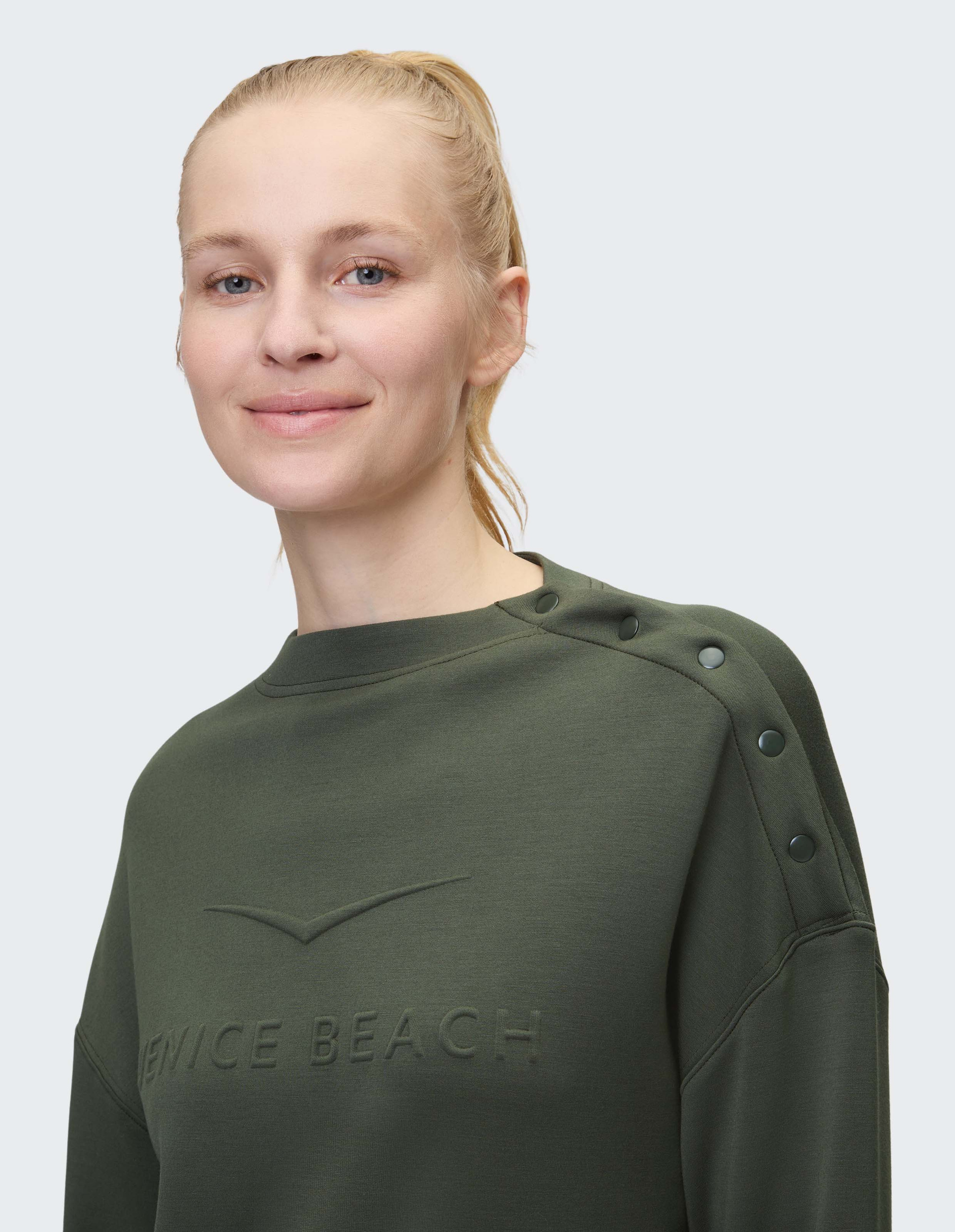 Eine lächelnde Person mit blondem Haar trägt ein olivgrünes Sweatshirt mit Knopfleiste auf der Schulter und dem Text "VENICE BEACH".