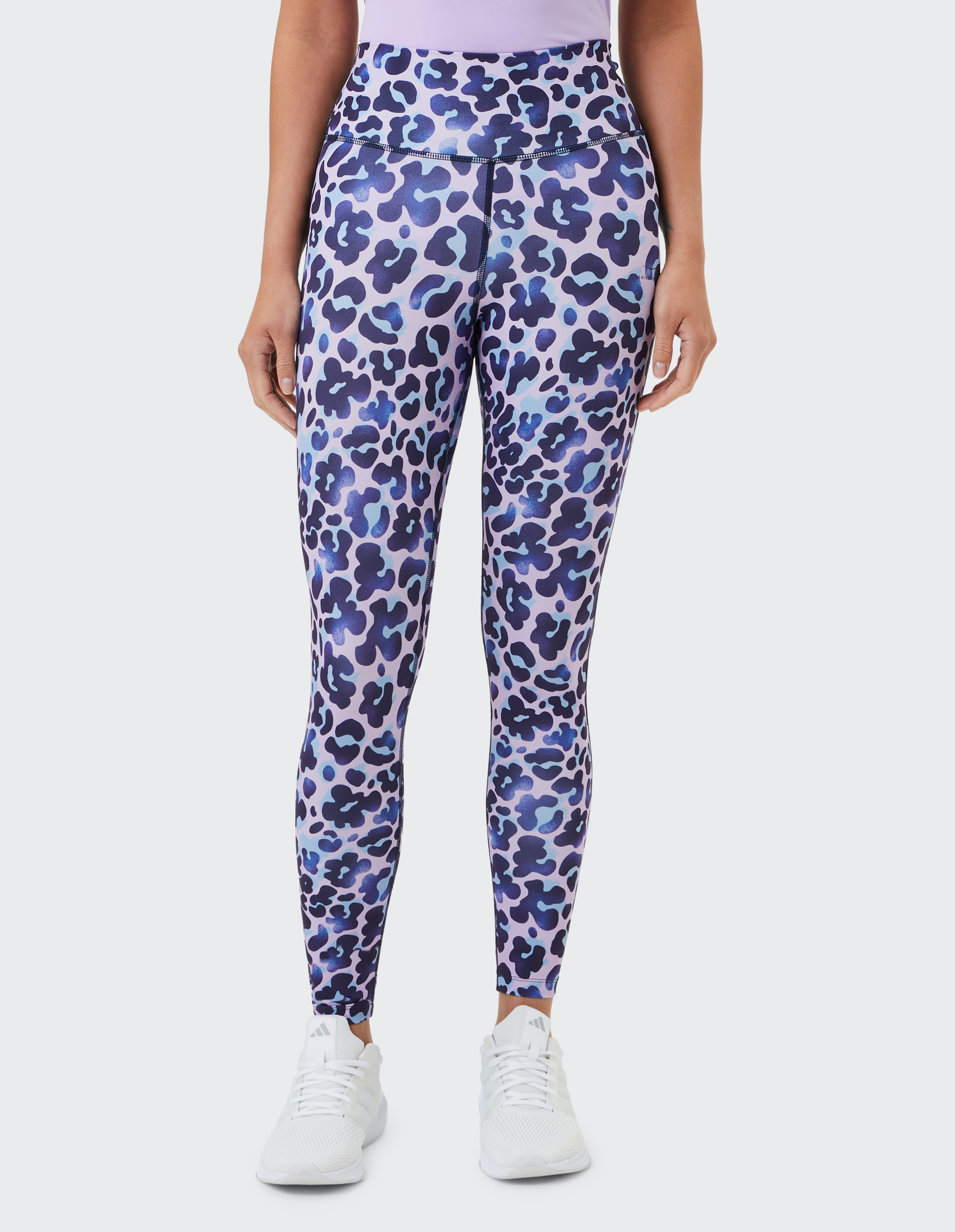 Frau trägt lilafarbene Leggings mit blau-schwarzem Leopardenmuster, dazu weiße Sportschuhe; Oberkörper nicht sichtbar.