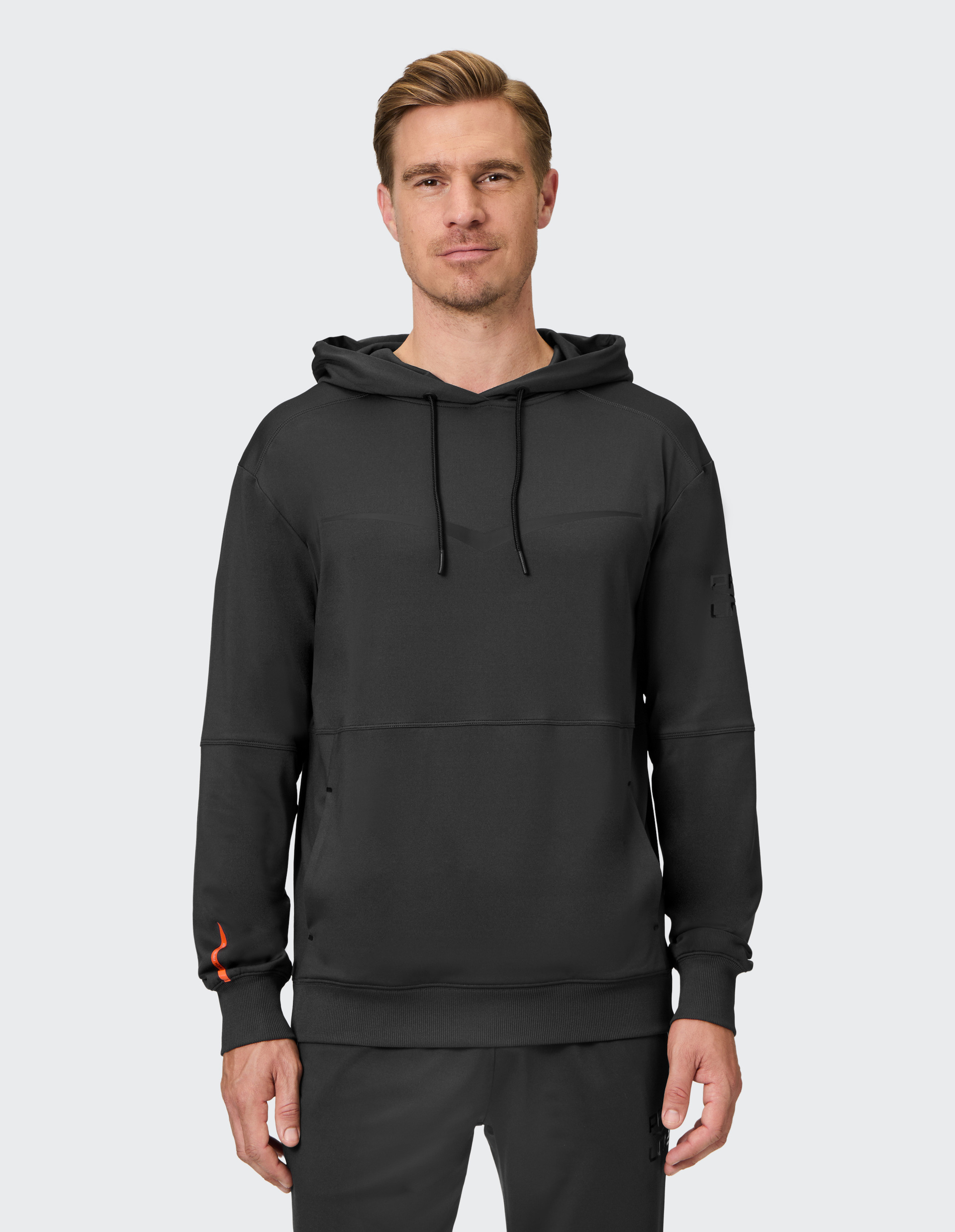Ein Mann trägt einen dunklen Hoodie mit Kapuze und passender Hose, vor einem hellgrauen Hintergrund stehend.