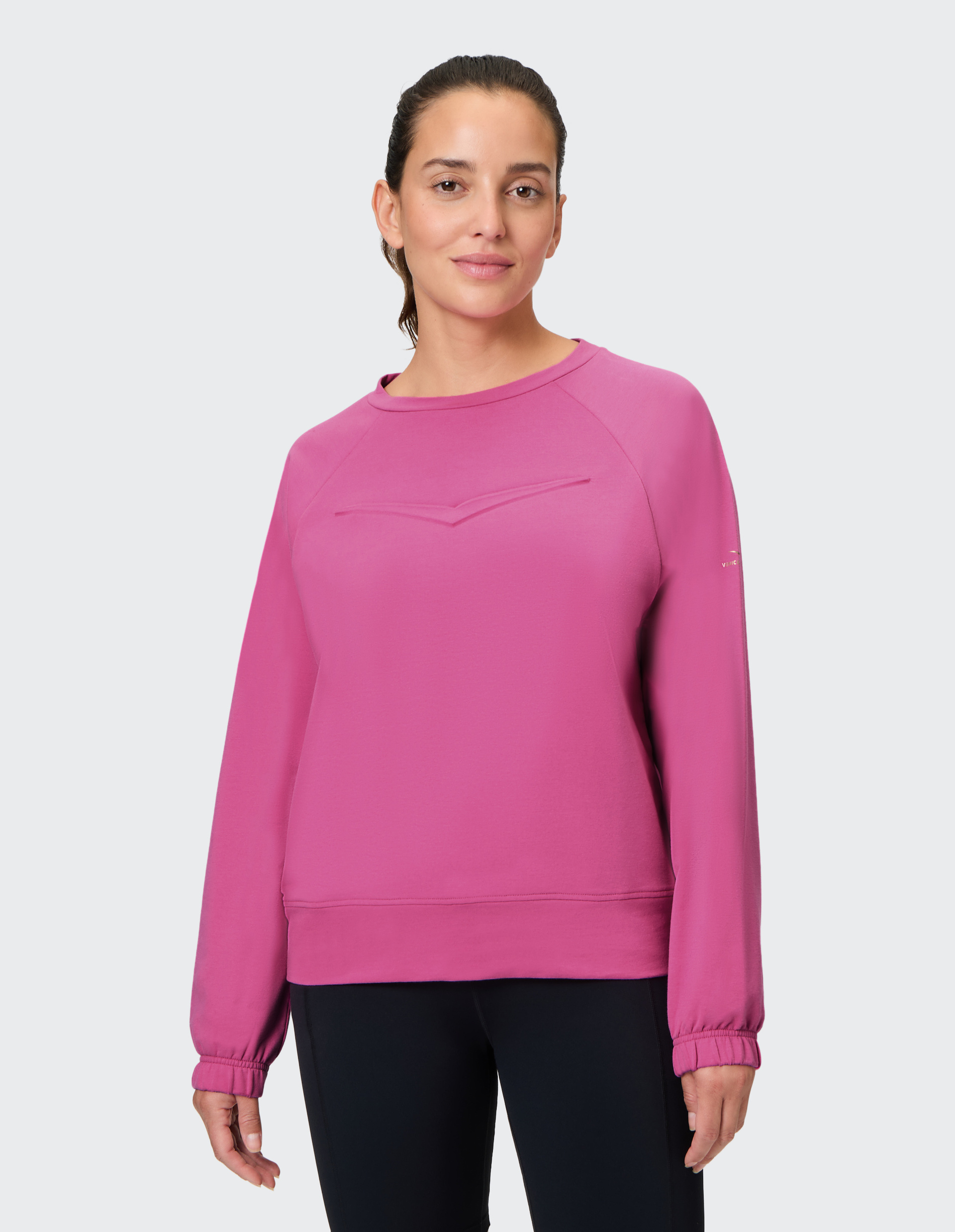 Frau in einem pinken Sweatshirt und schwarzen Leggings steht lächelnd vor einem neutralen Hintergrund.