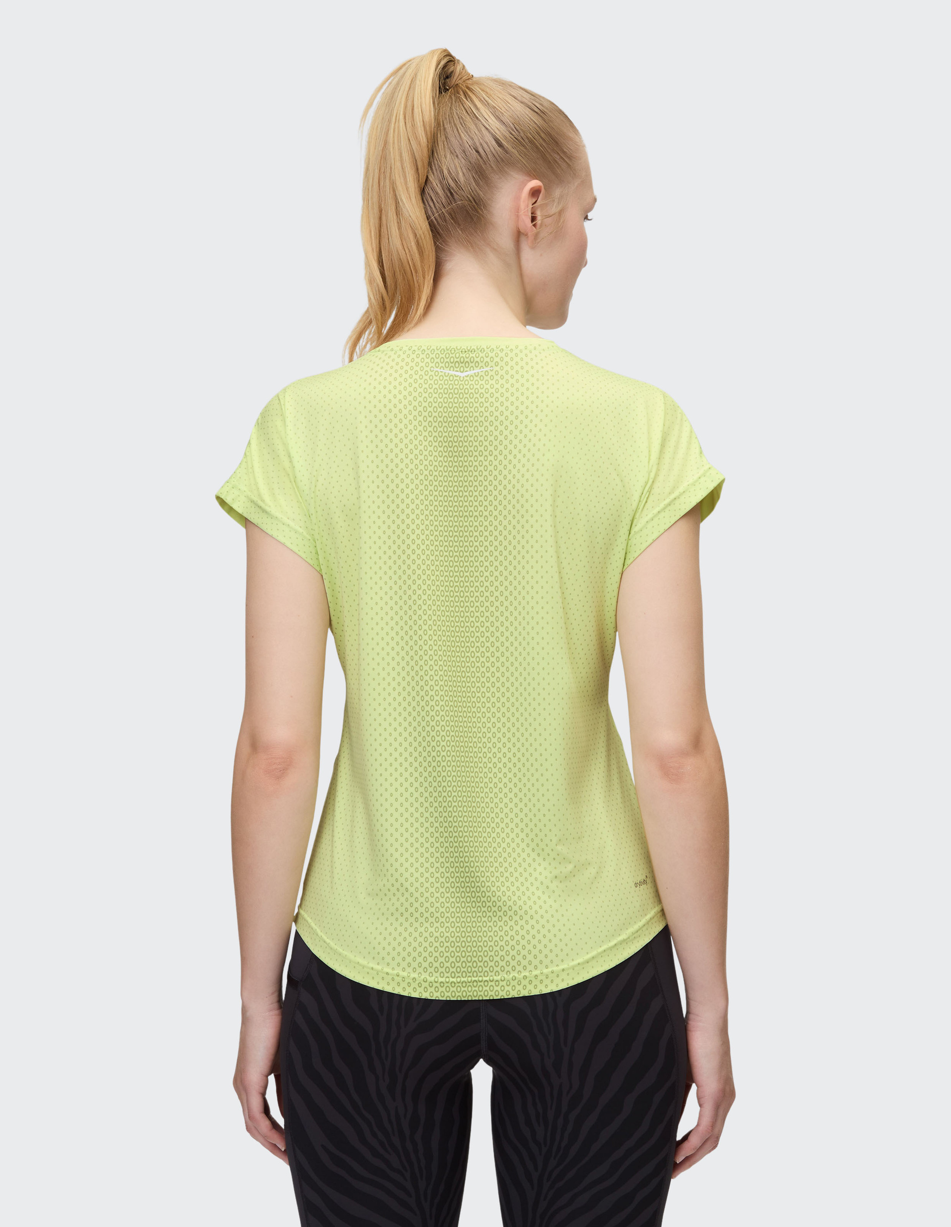 Eine Person mit blondem Pferdeschwanz trägt ein gelbes Mesh-Shirt und schwarze gemusterte Leggings, von hinten fotografiert.
