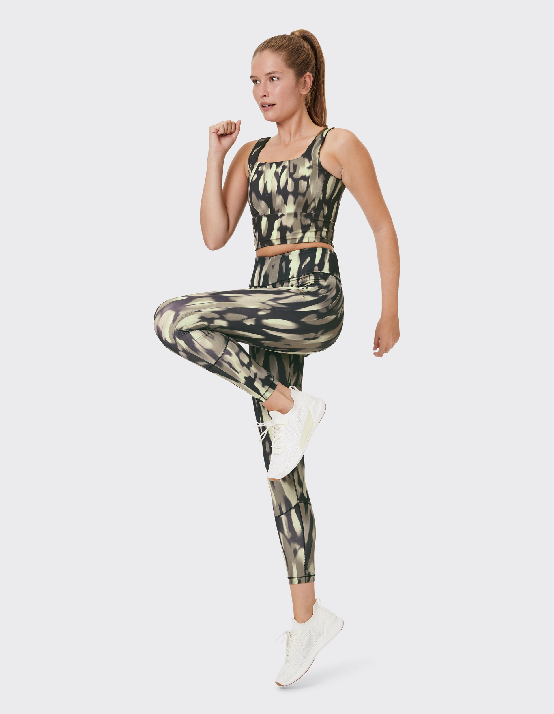 Frau in gemusterten Sportbekleidung macht Fitnessübung, hebt Knie an, trägt weiße Turnschuhe und hat lange Haare im Zopf.